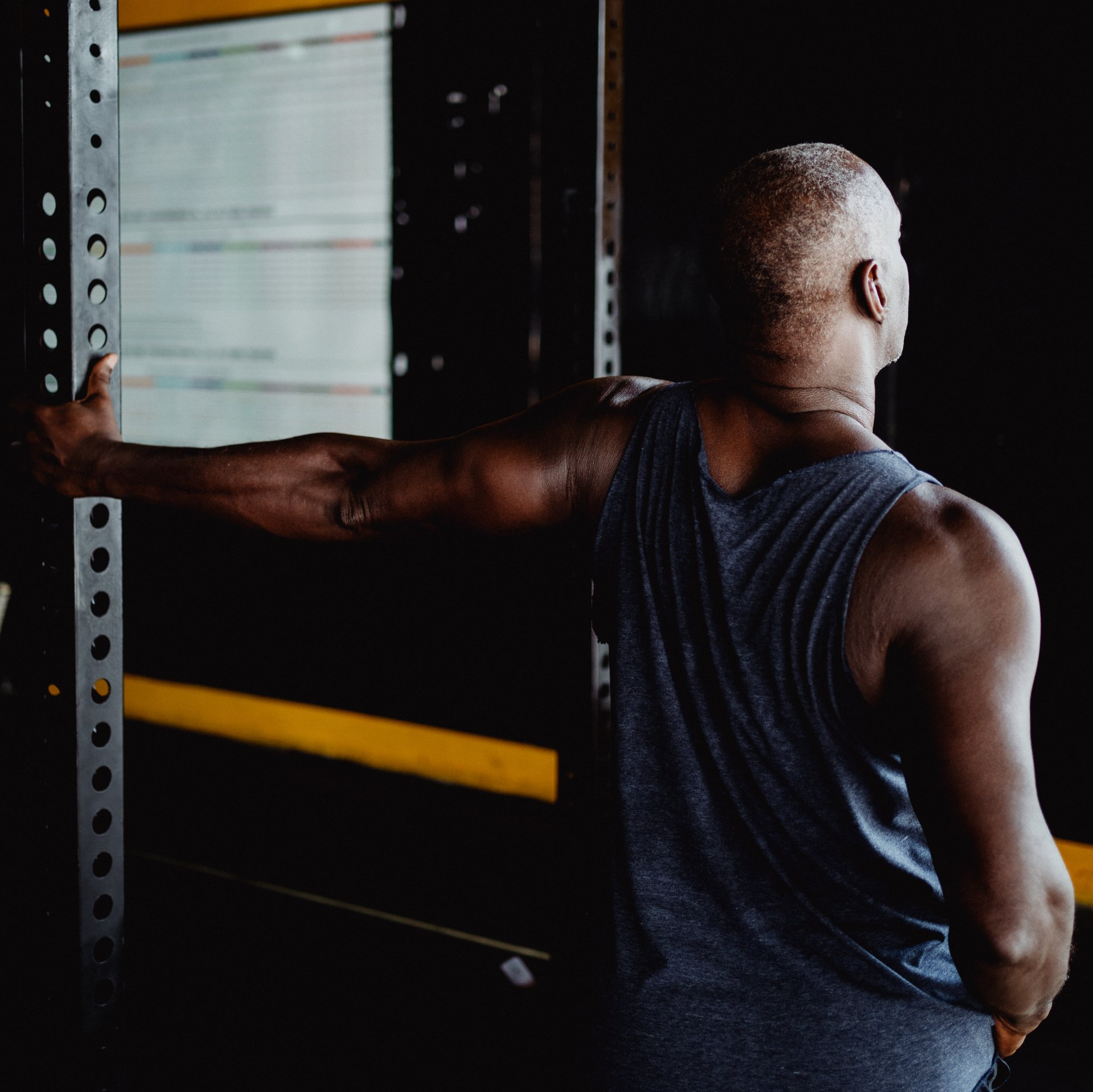 A man in a tank top performing a rotator cuff stretch in a gym, focusing on shoulder flexibility and mobility exercises