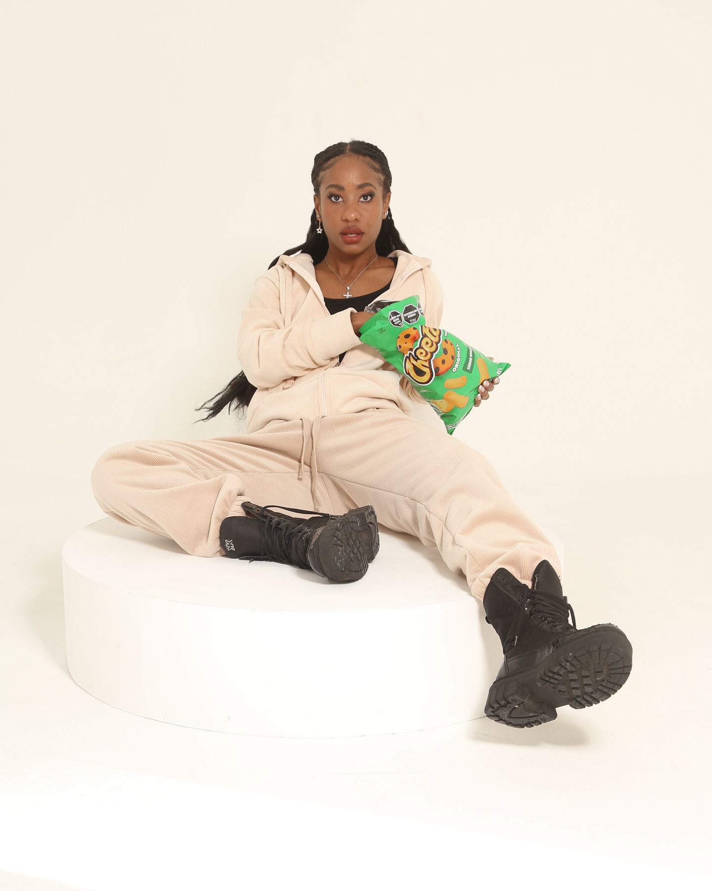 Mujer con chándal color canela y botas negras sosteniendo una bolsa de patatas fritas, de pie sobre una superficie blanca.