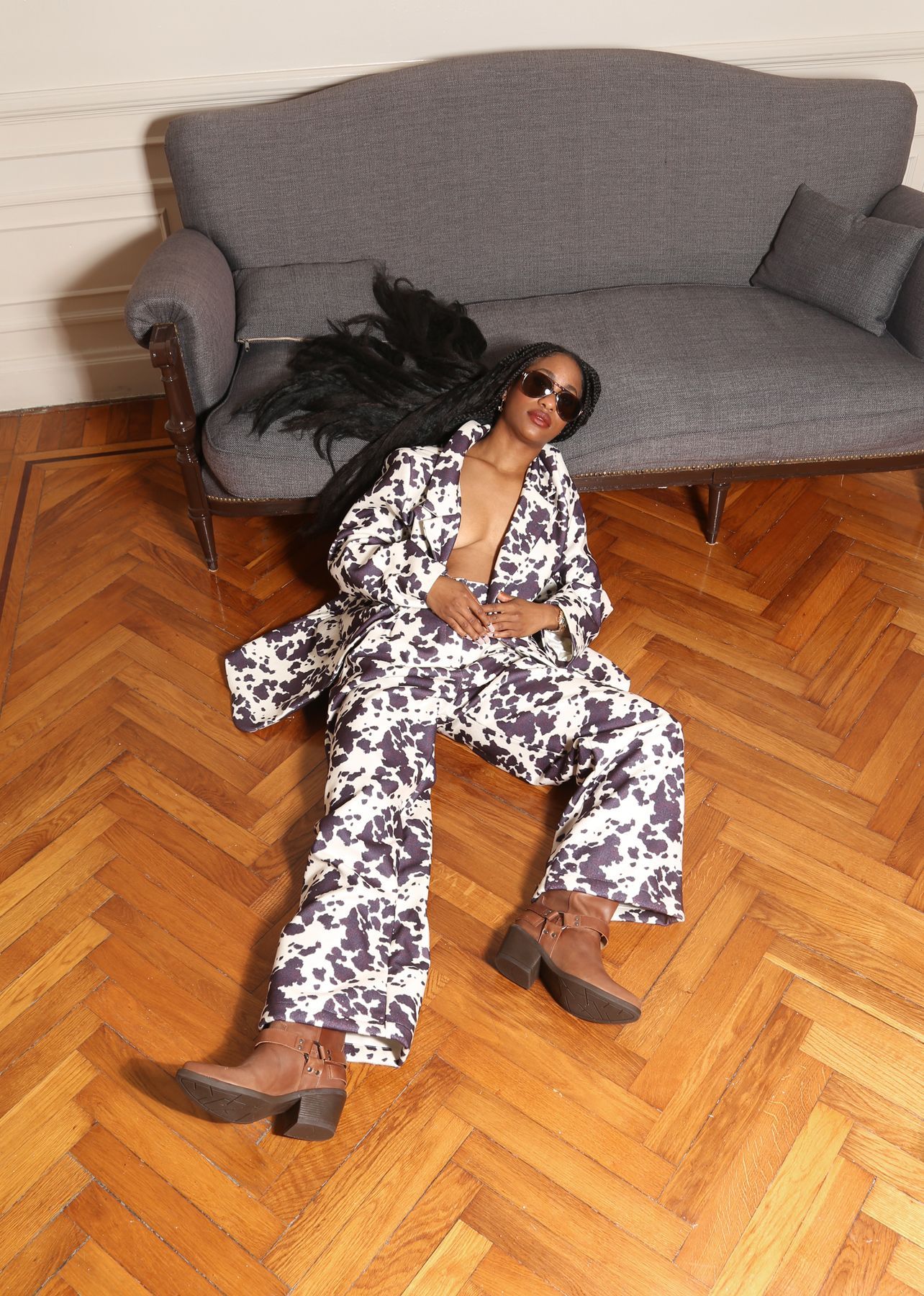 Una persona con traje estampado y gafas de sol descansa sobre un suelo de madera cerca de un sofá gris.