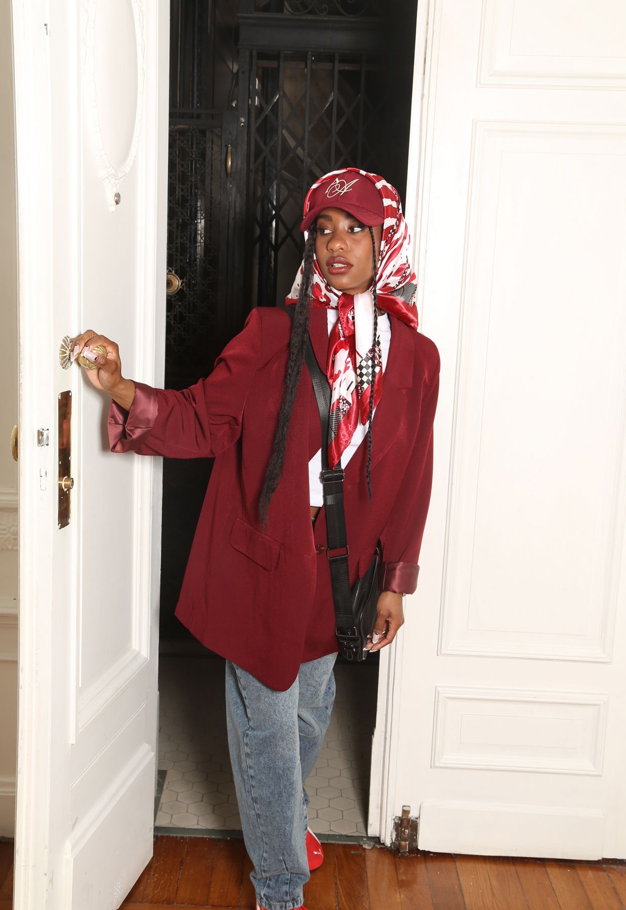 Mujer con blazer burdeos, vaqueros, pañuelo en la cabeza y zapatillas rojas posa junto a una puerta blanca.