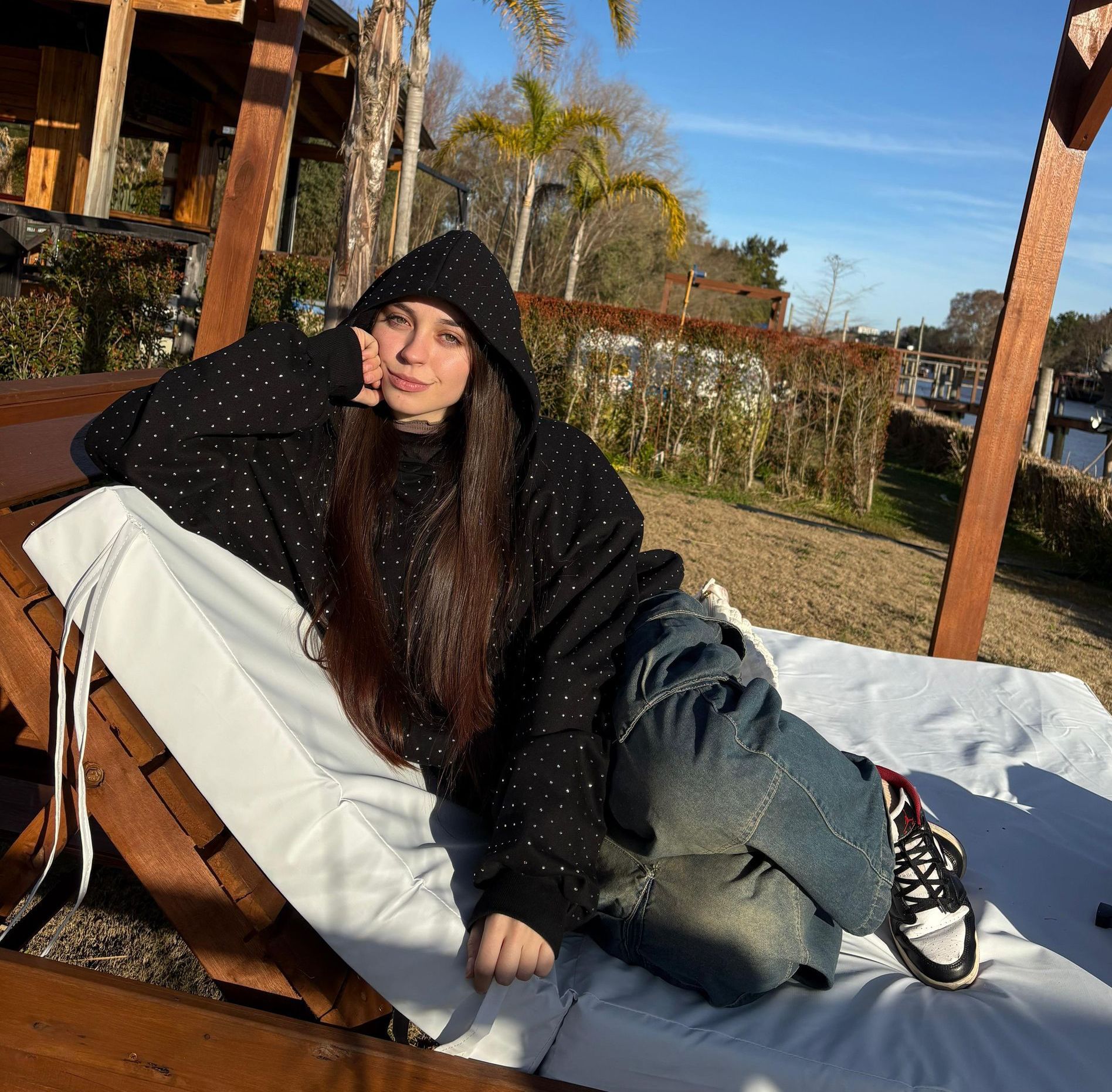 Una mujer con sudadera negra con capucha está sentada en una tumbona de madera en el exterior, cerca del agua.