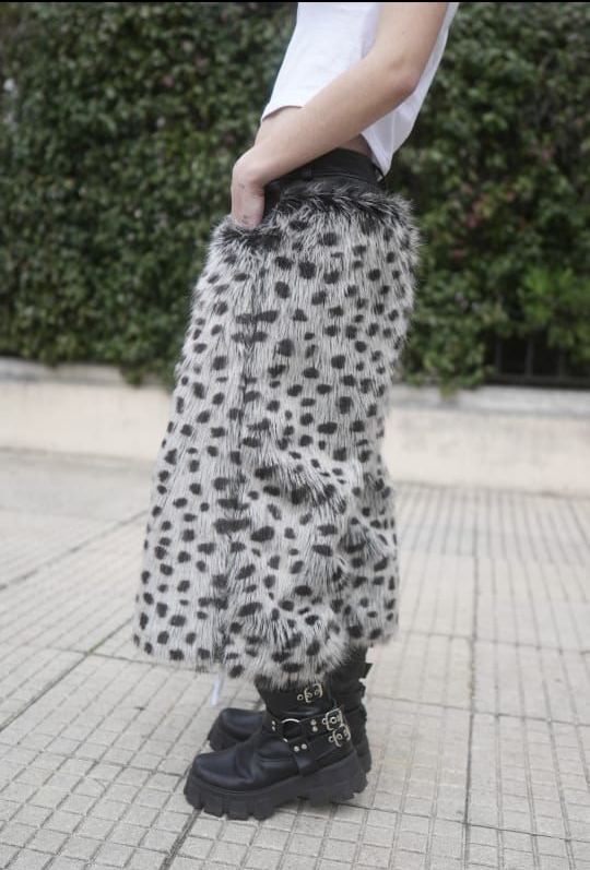 Mujer con camiseta blanca y pantalones con estampado animal, apoyada contra una columna blanca, sosteniendo un objeto blanco.