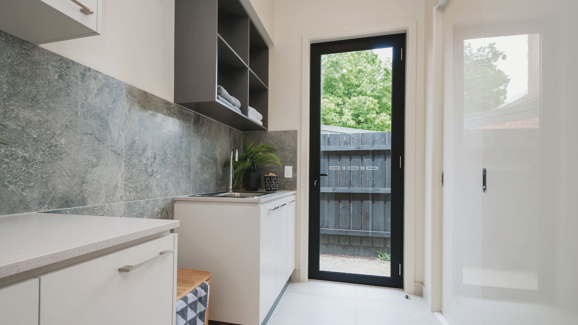 Modern laundry room with white cabinets, grey backsplash, and black-framed door leading to a backyard — K & J Glass in Bowraville, NSW