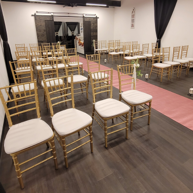 Gold chairs with white cushions arranged for a ceremony, pink aisle runner, dark wood floor.