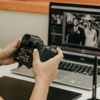 Person holding a camera, reviewing settings while a wedding photo is edited on a laptop in the background.