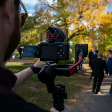 A person filming a wedding procession with a camera on a stabilizer. The background features autumn trees and people.