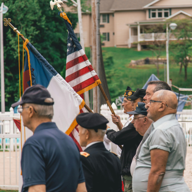 Veterans stand with flags at an outdoor ceremony, with houses in the background.