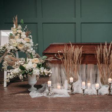 Coffin with floral arrangements, candles, and a white draped fabric in front of a dark green wall.