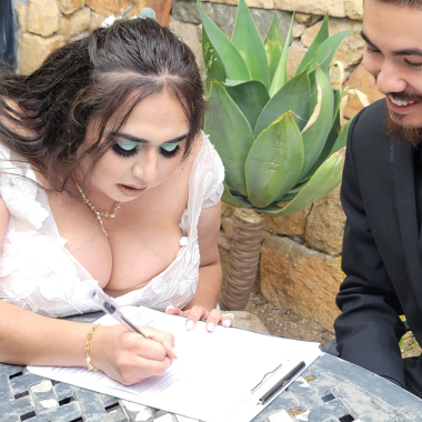 Bride signing documents outdoors, groom smiles beside her.