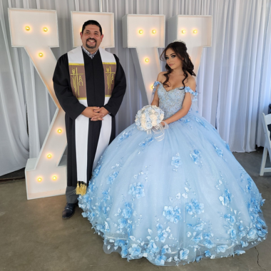 A woman in a light blue ballgown and a man in clergy attire stand before a large light-up 