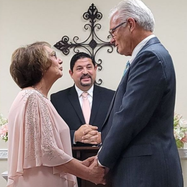 Older couple holding hands at a wedding ceremony; officiant smiles in front of a cross.