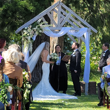 Wedding ceremony: bride, groom, officiant under decorated arbor, guests seated outdoors.