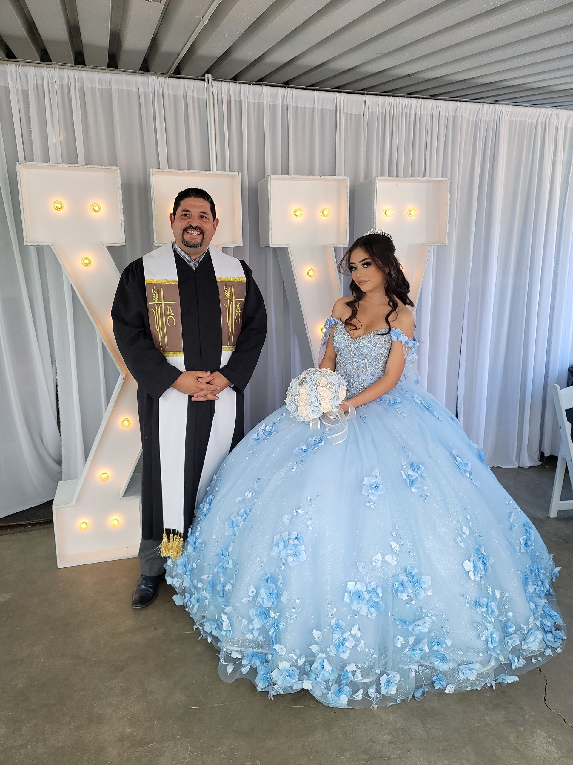 Quincenera in blue dress with white bouquet stands in front of Lit
