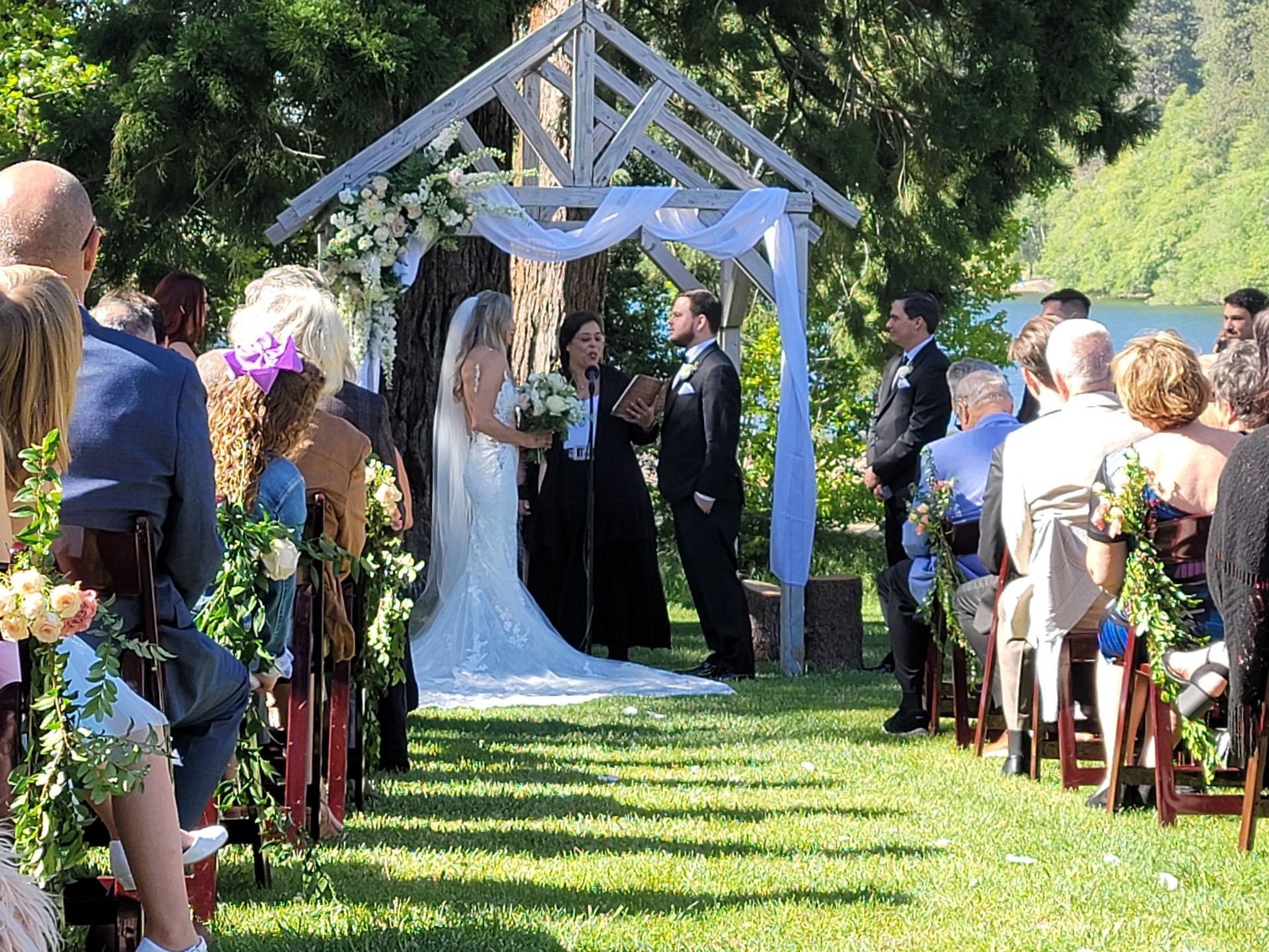 Wedding ceremony by a lake; bride and groom stand under a white arch, surrounded by guests on a sunny day.