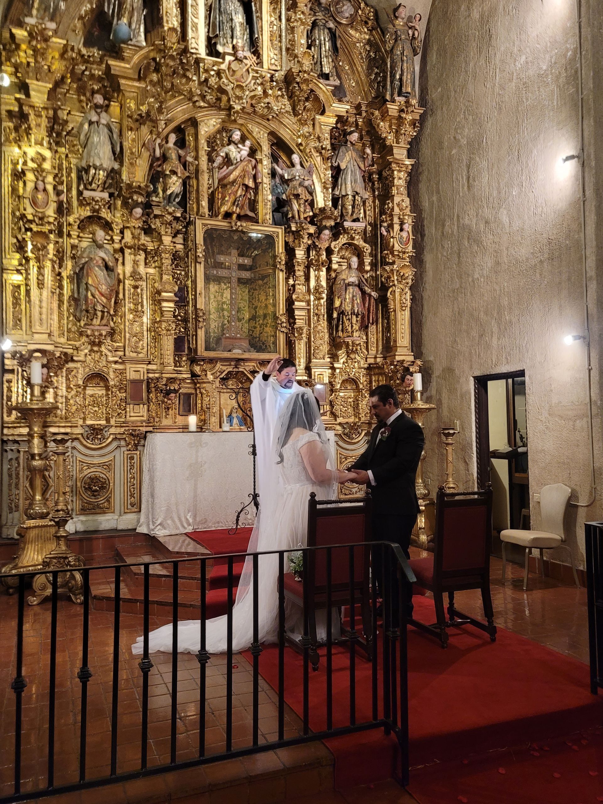 Wedding ceremony in a church with ornate gold altar. Bride and groom at the altar, red carpet, dark attire.