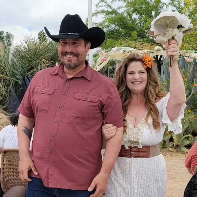 Newly married couple: man in cowboy hat, woman in white dress holding bouquet, smiling, outdoors.