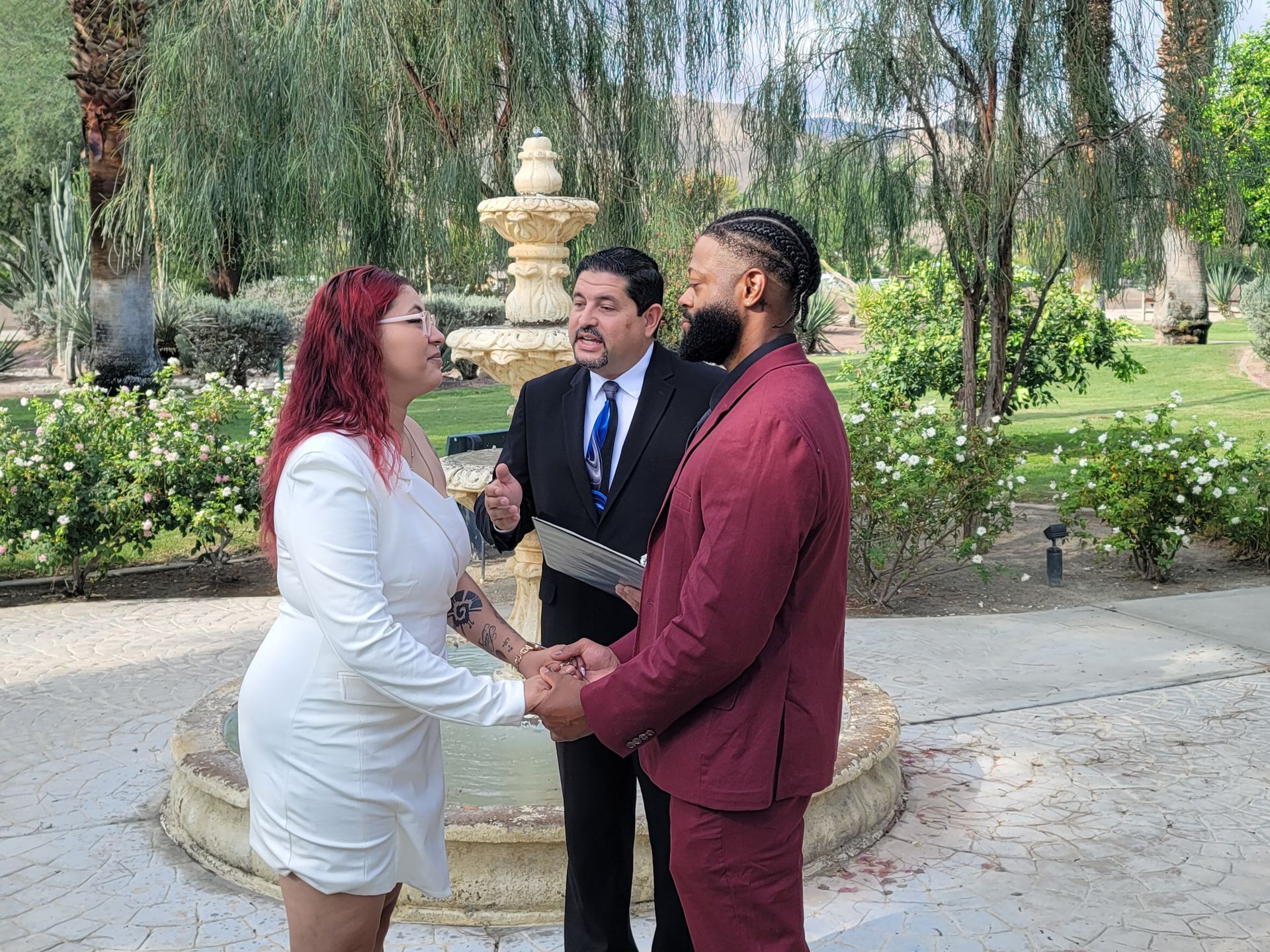 Couple holding hands during a wedding ceremony outdoors. A man in a suit officiates.