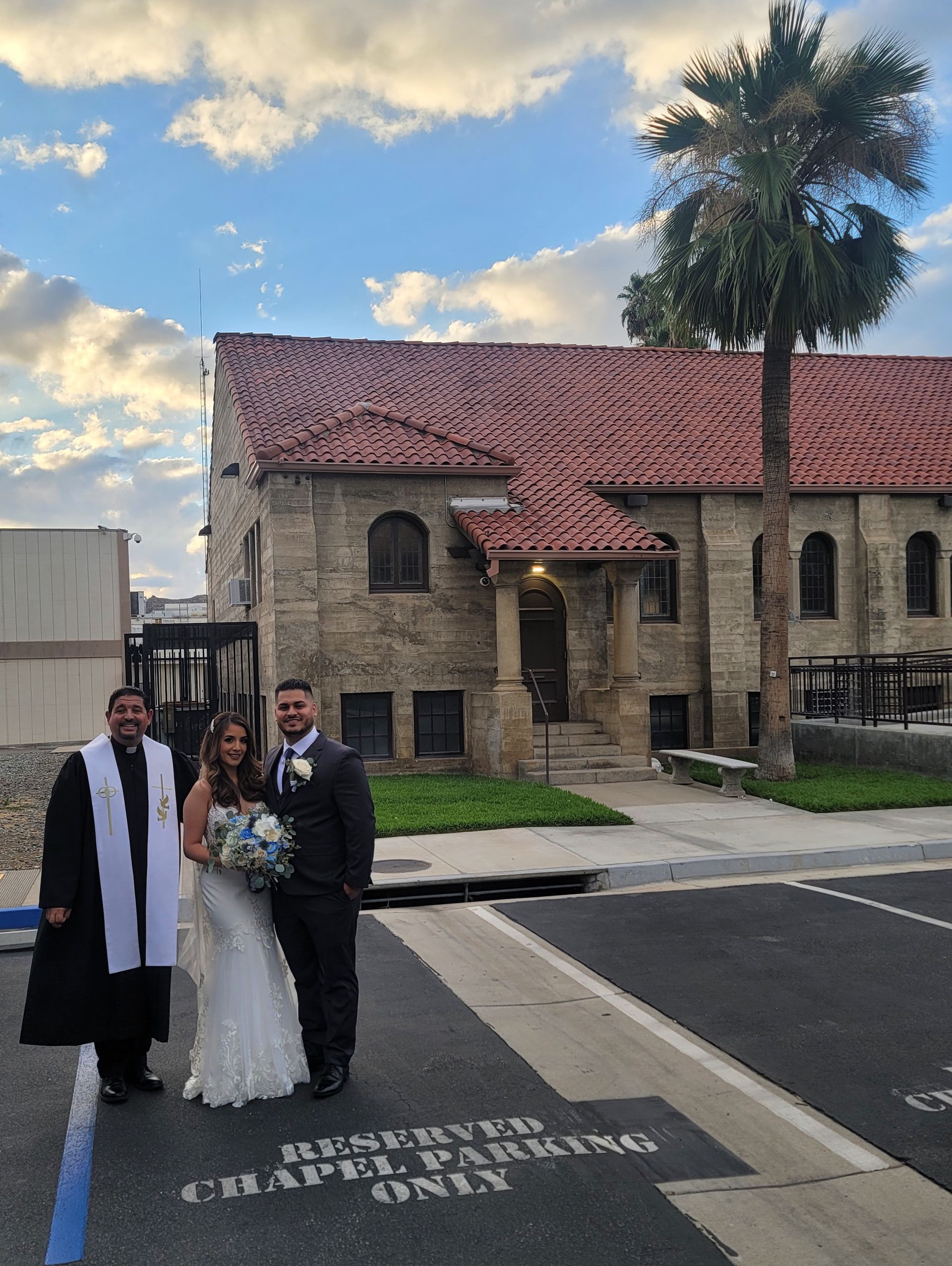Bride and groom with officiant in front of a stone chapel with red-tiled roof and palm tree.