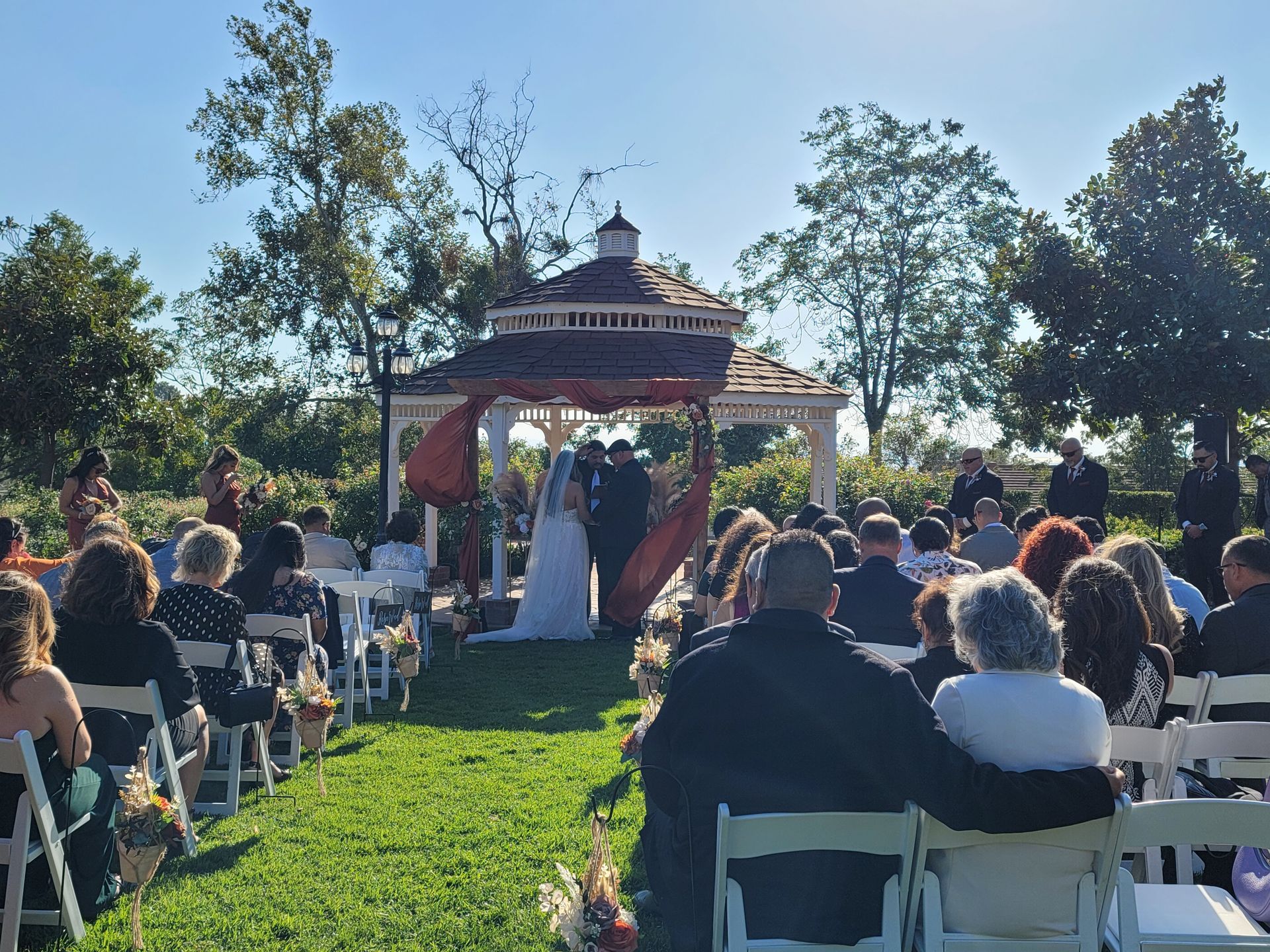 Wedding ceremony outdoors: bride and groom under gazebo, guests seated on lawn. Bright sunlight.
