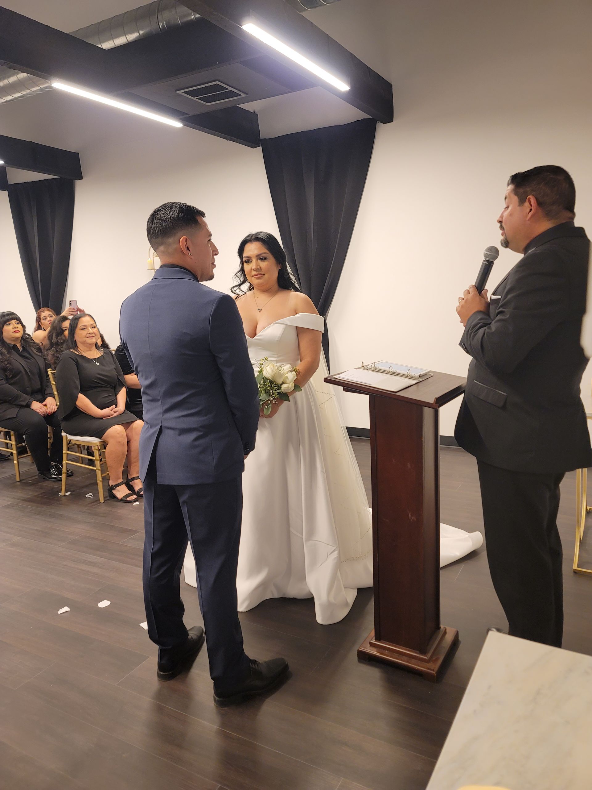 Couple facing officiant at altar. Bride in white gown, groom in blue suit, officiant speaking. Guests seated.