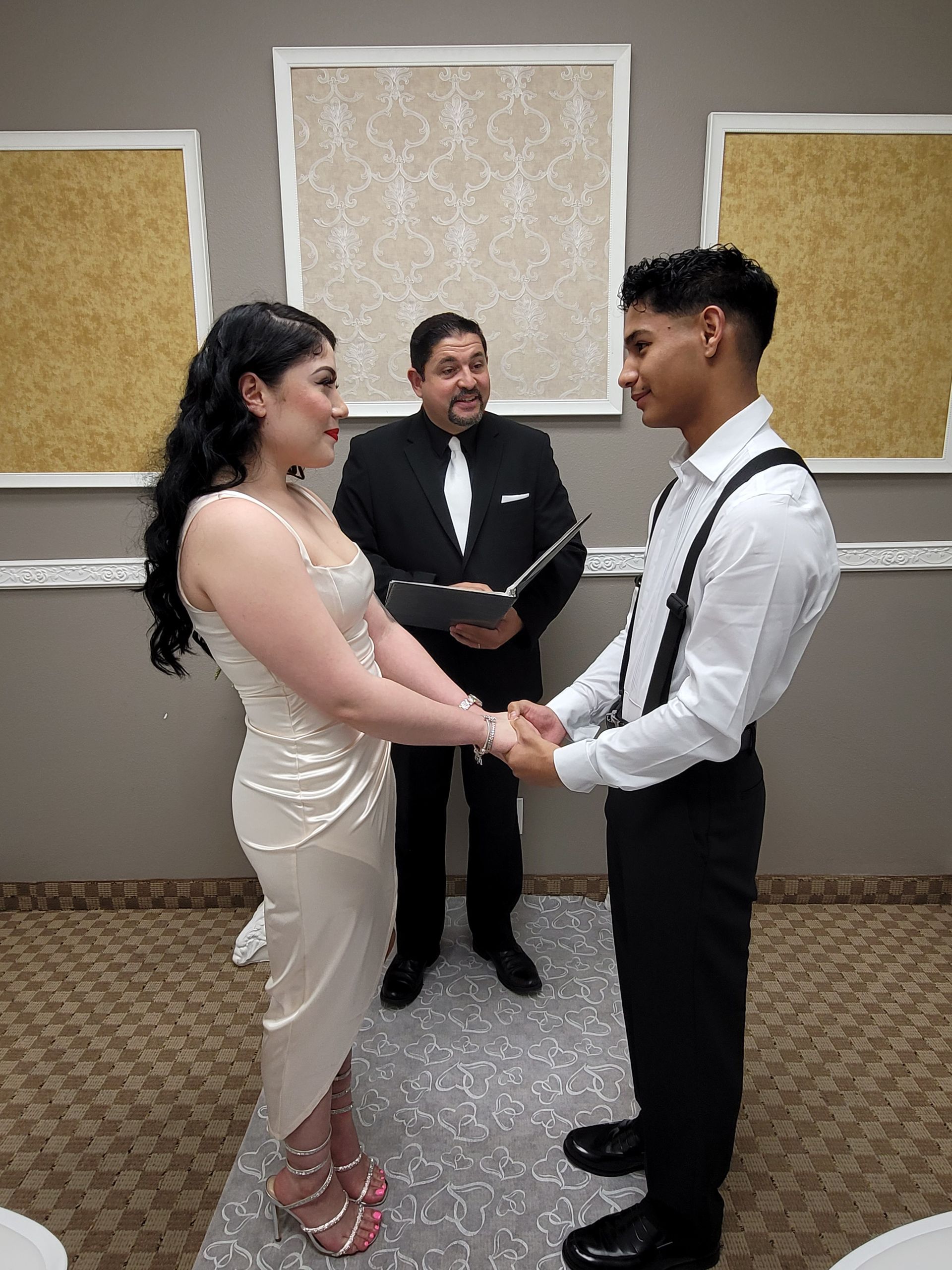 Couple holding hands at wedding ceremony, officiant behind them. Beige dress, black pants, gold decor.