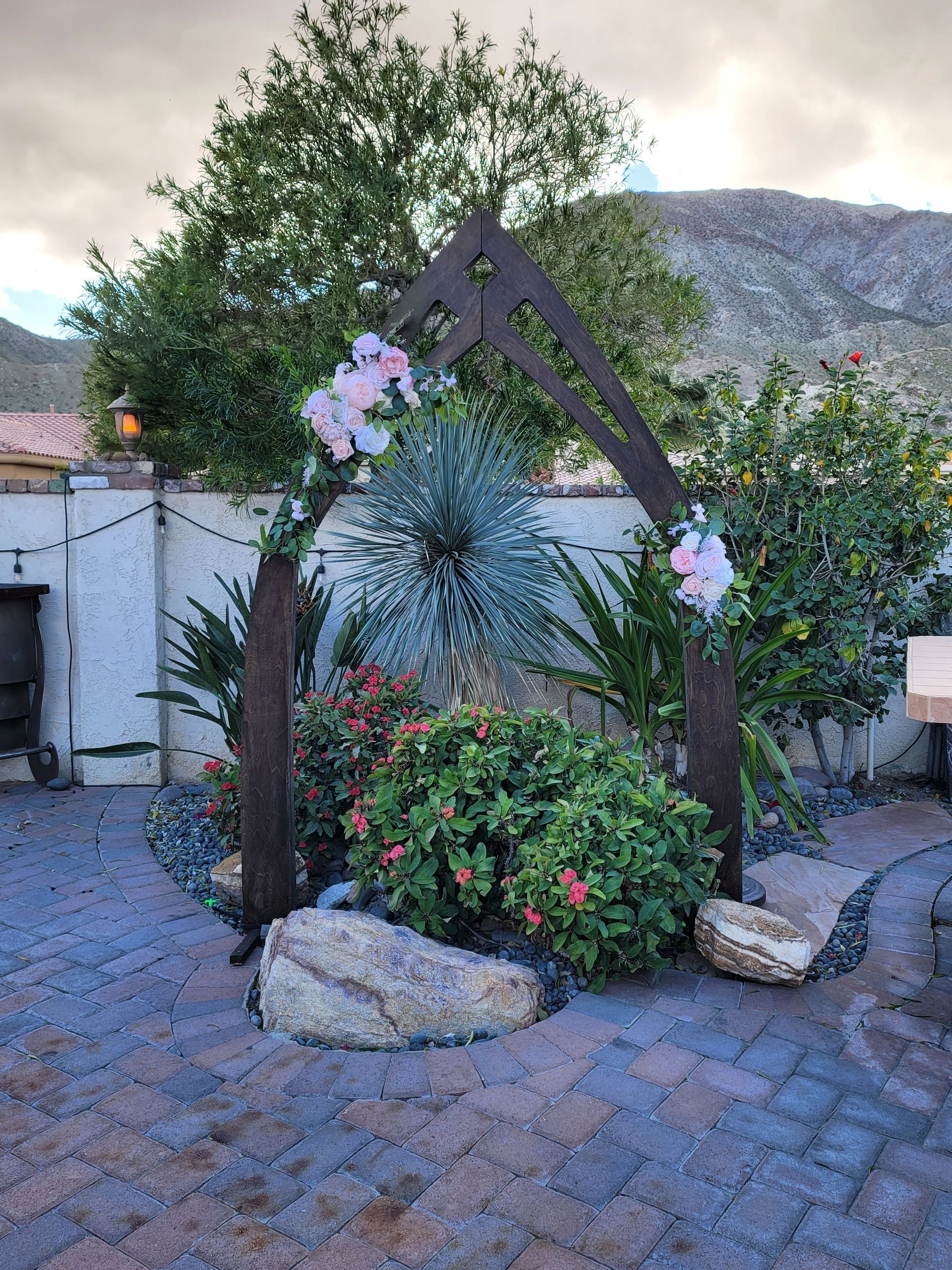 Arched metal structure decorated with pink flowers and greenery, set in a garden with brick pathway.