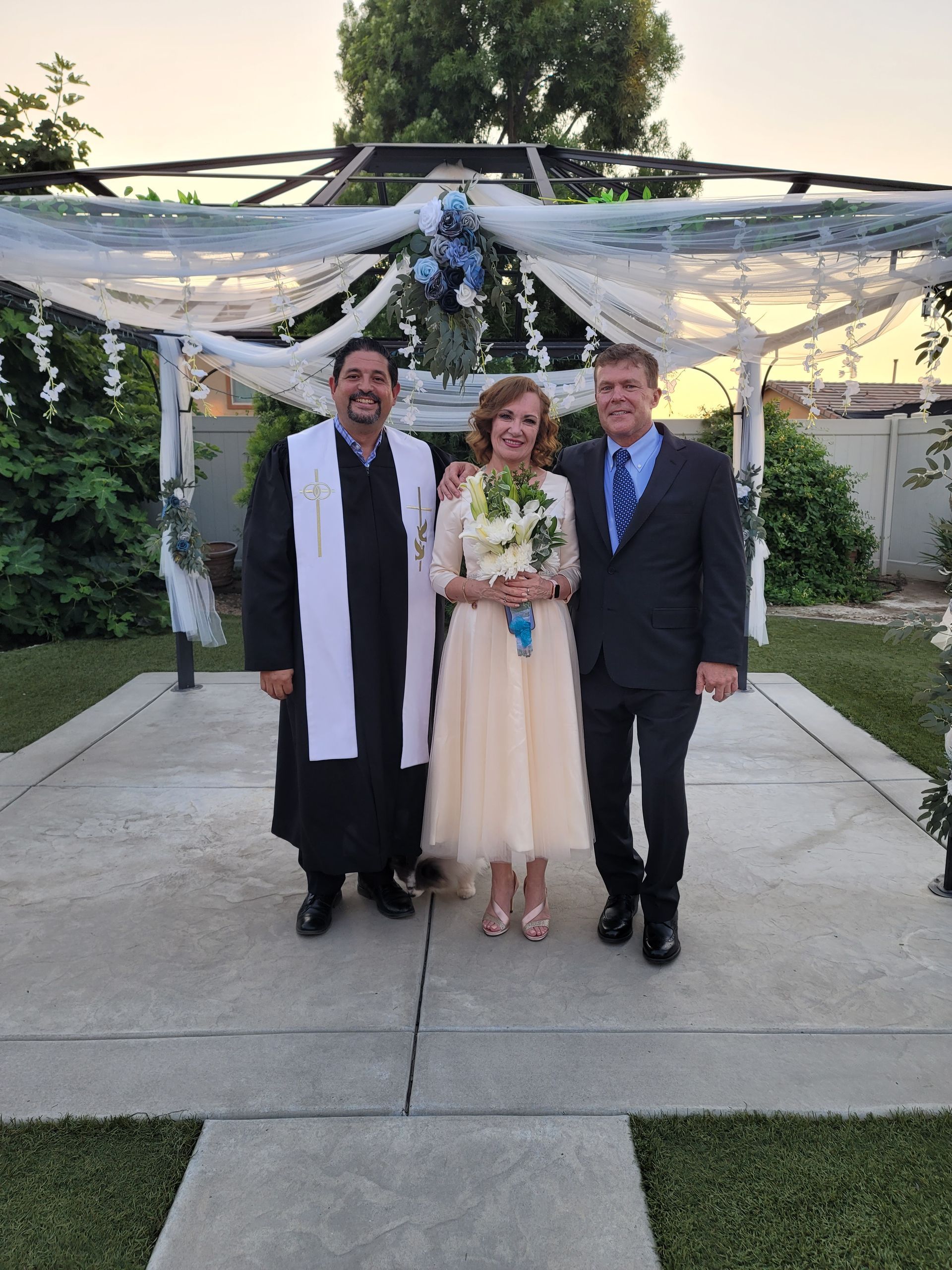 Couple and officiant pose for a wedding photo under a decorated trellis in an outdoor setting.