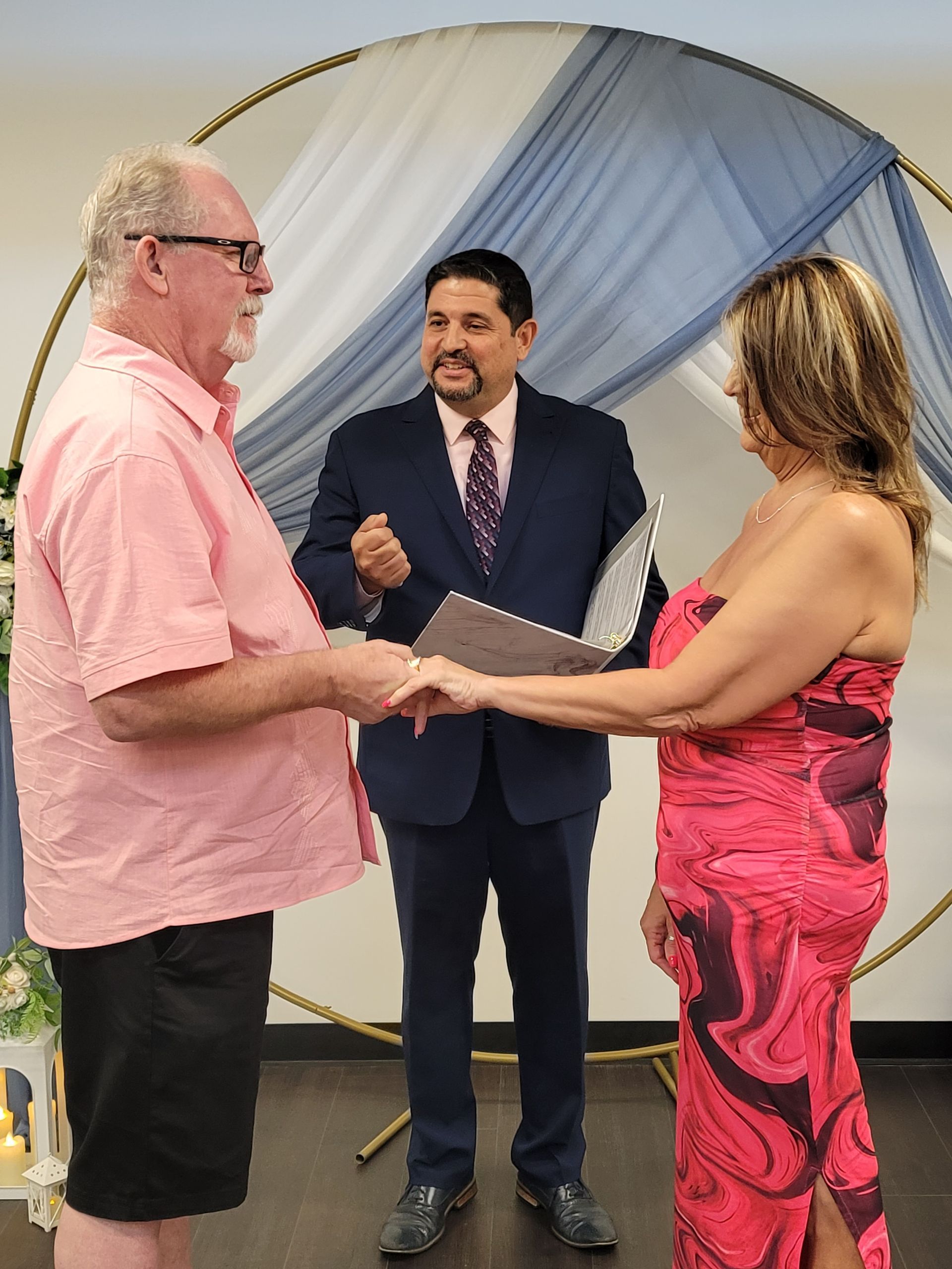 Couple holding hands during a wedding ceremony; officiant in suit; blue and gold backdrop.
