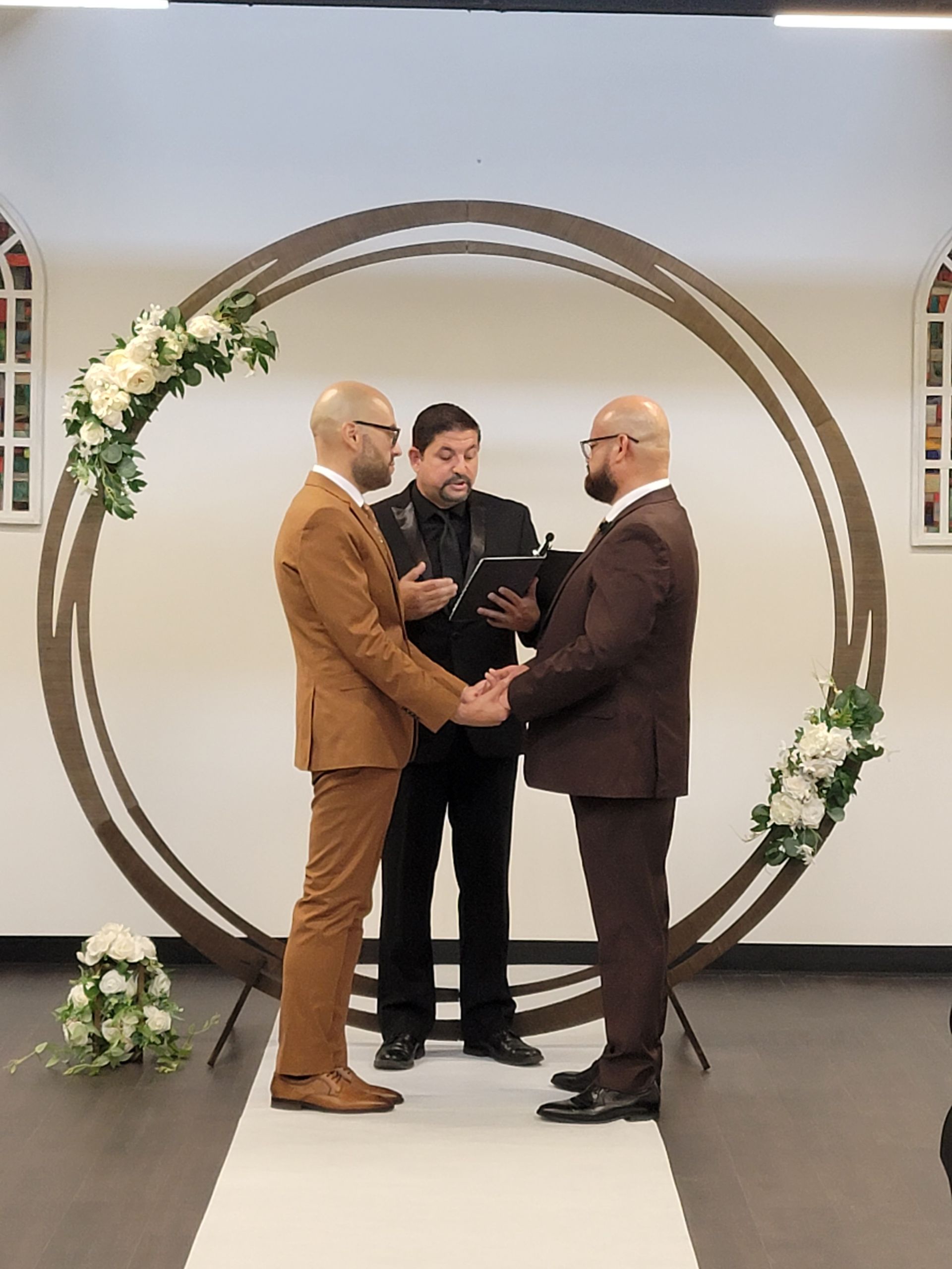Two men holding hands during wedding ceremony, facing officiant under floral archway.