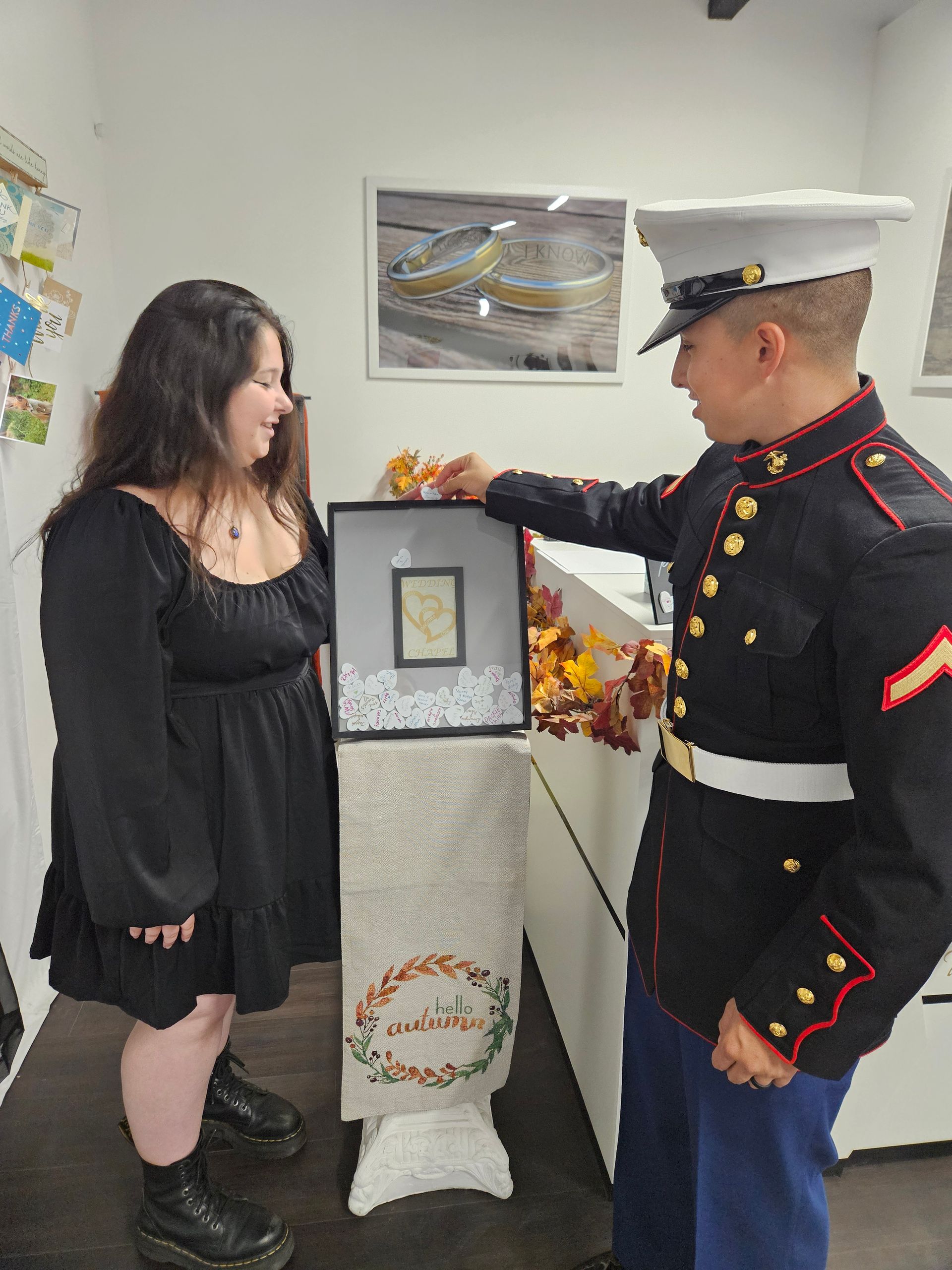 A Marine in uniform presents a framed item to a woman in a black dress; they are smiling in an office.