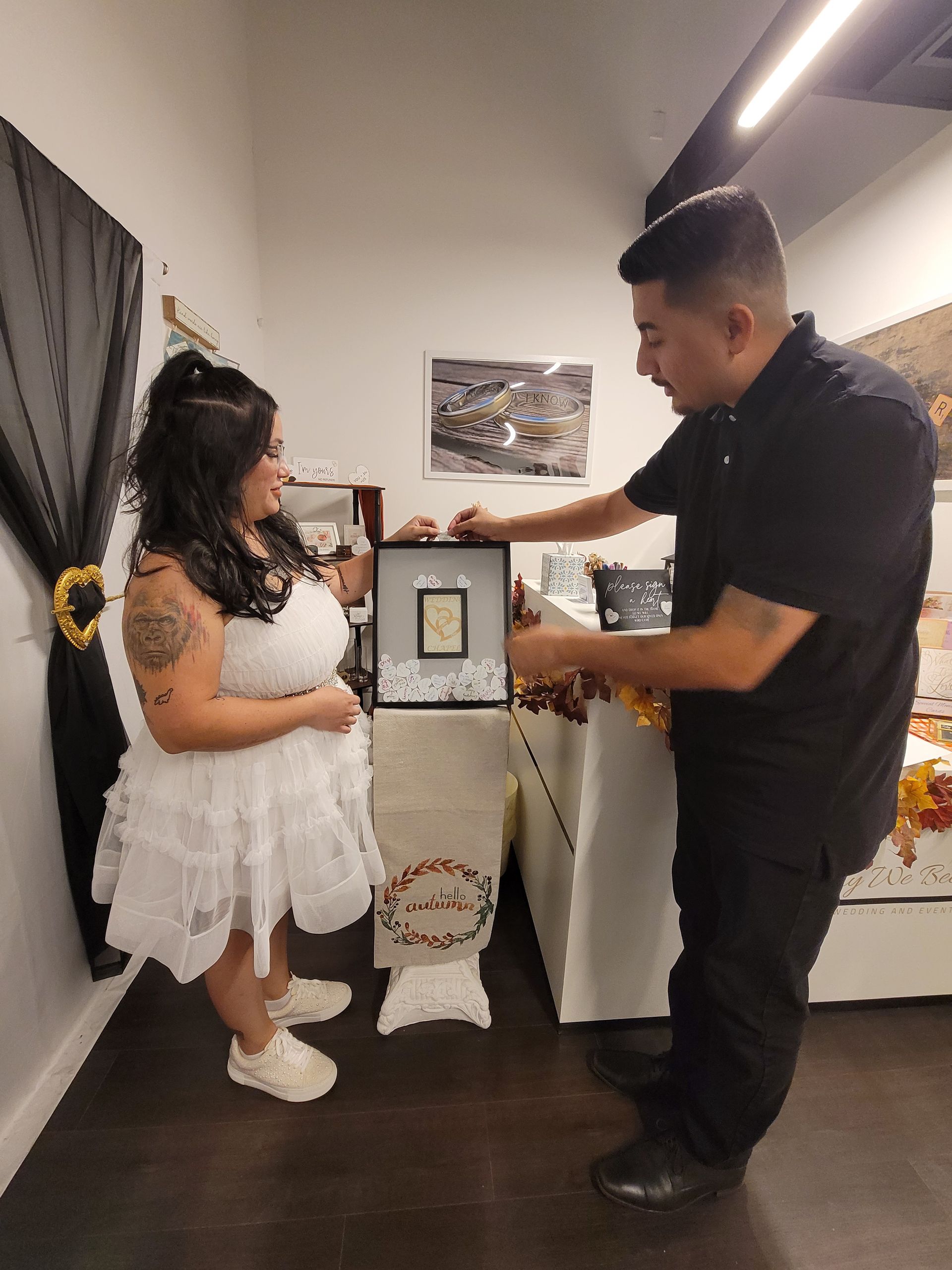 Woman in white dress, man in black shirt, looking at framed artwork on display table.