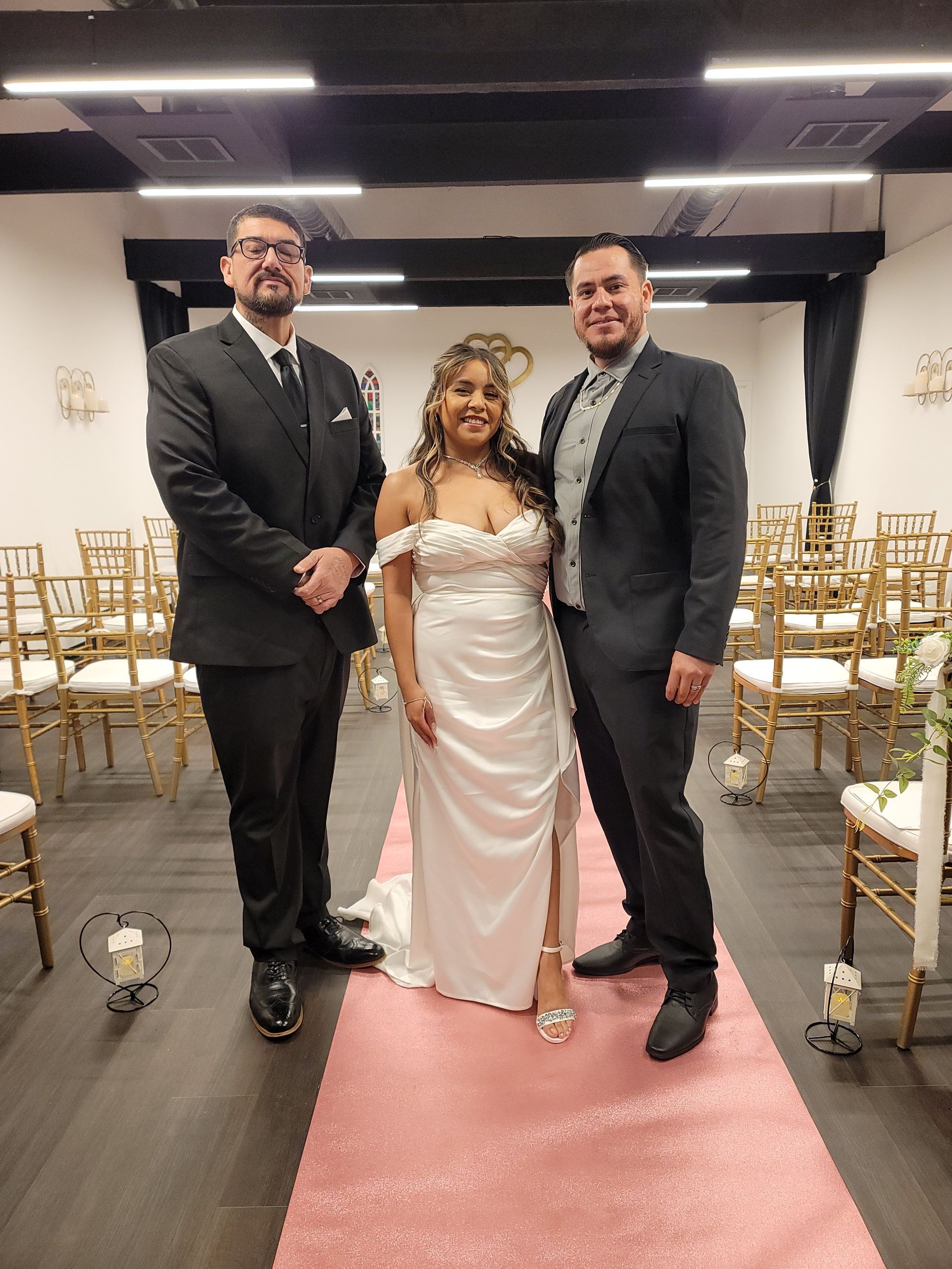 Bride with two men posing on a pink carpet aisle. Indoor wedding venue with gold chairs.