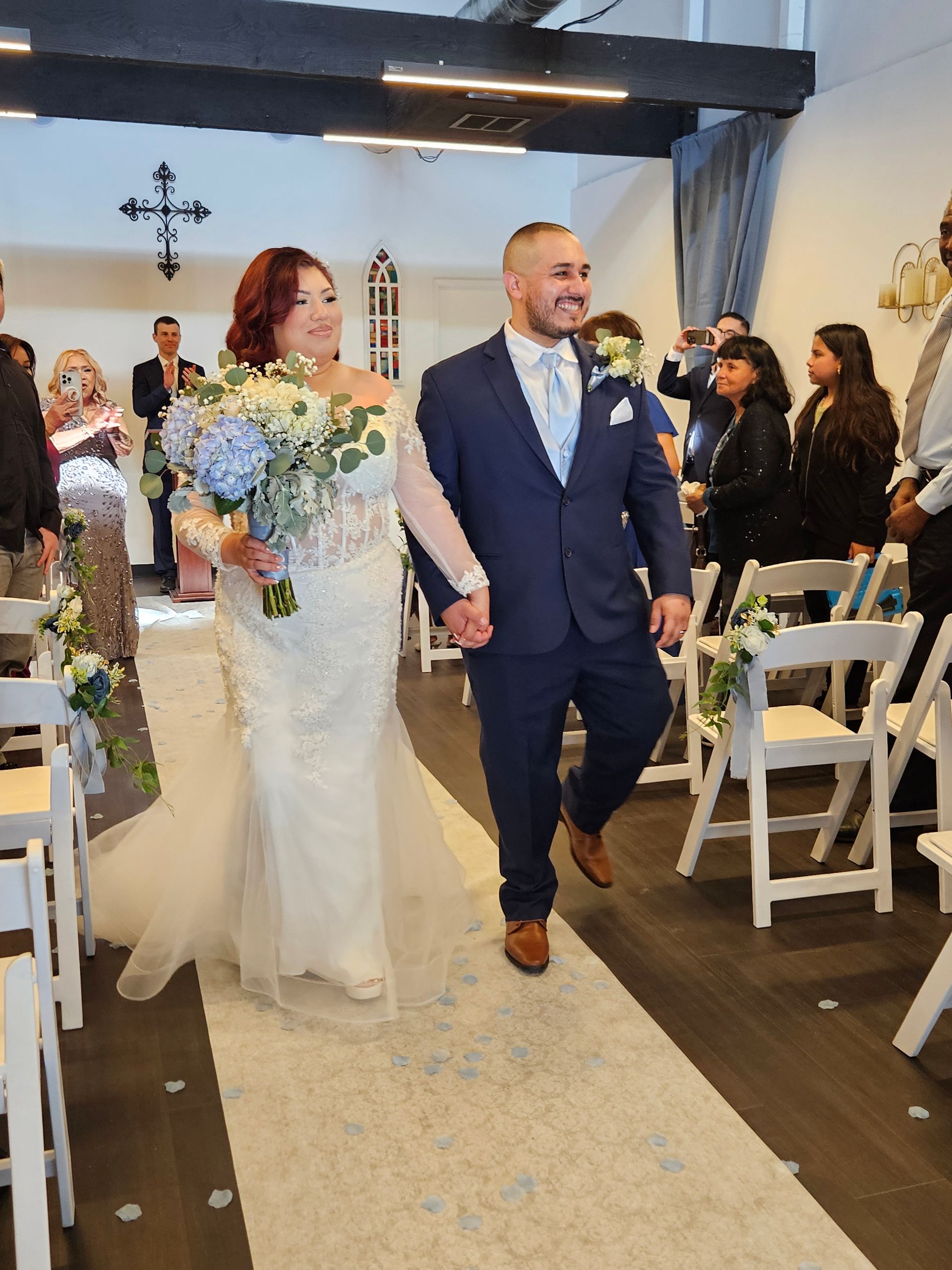 Newlyweds walk down the aisle, bride in white dress with flowers, groom in blue suit, at a wedding.