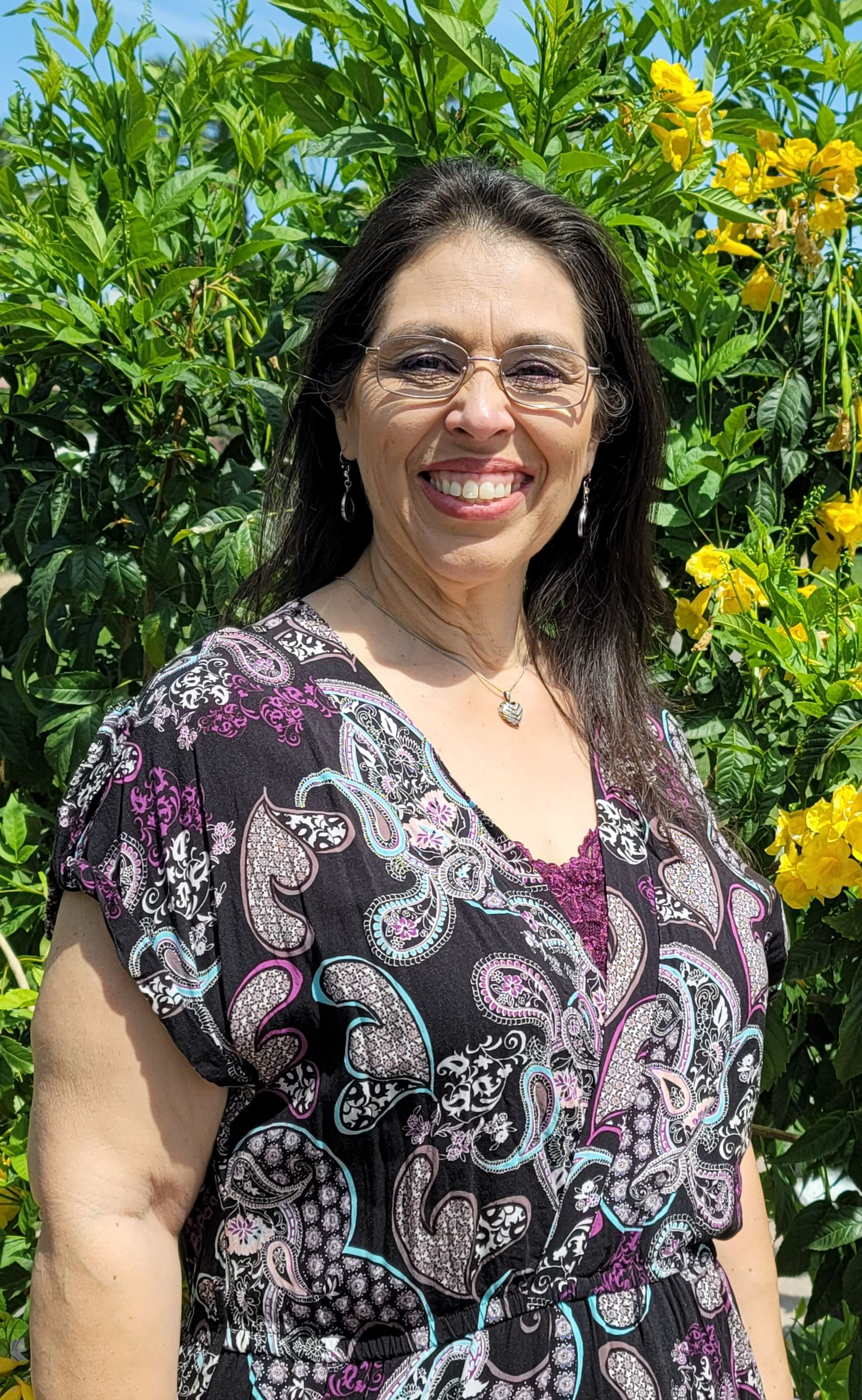 Woman with glasses smiles, wearing a patterned dress, standing outside with flowers.