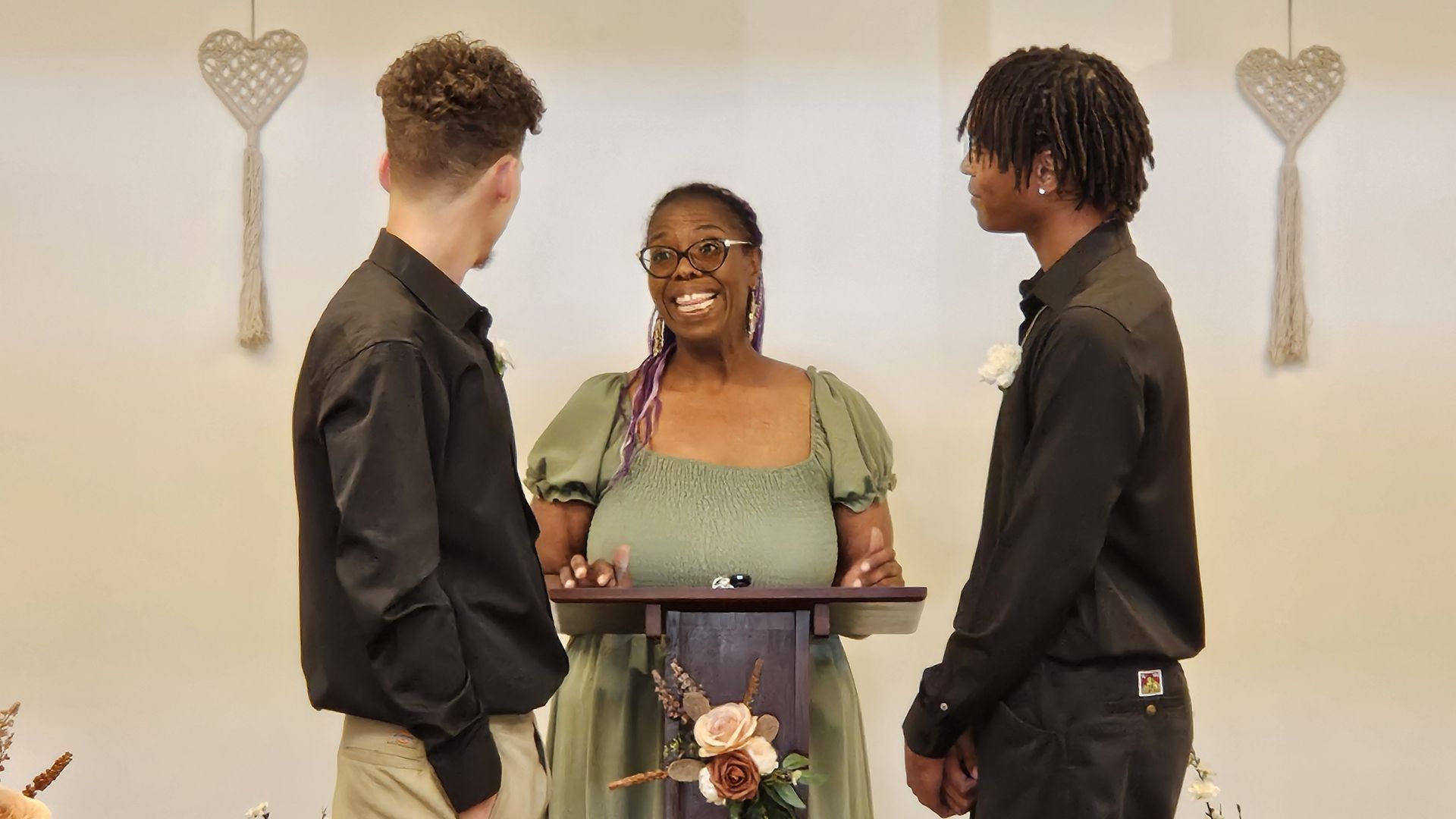 Two men at a wedding ceremony, facing a smiling officiant. They stand indoors, dressed in black shirts.