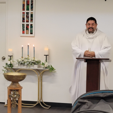 A priest in white robes at a podium, candles on a table, and a baptismal font.