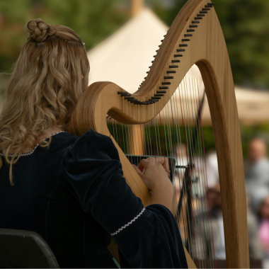 Woman plays a wooden harp outdoors, wearing a dark blue dress with a white-trimmed collar.