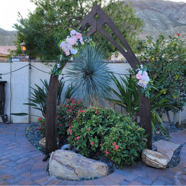 Wooden wedding arch with pink flowers, set in a garden with desert plants and rocks.
