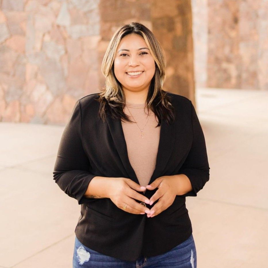 Woman in black blazer smiles, hands clasped. Standing outdoors, backdrop of stone wall.