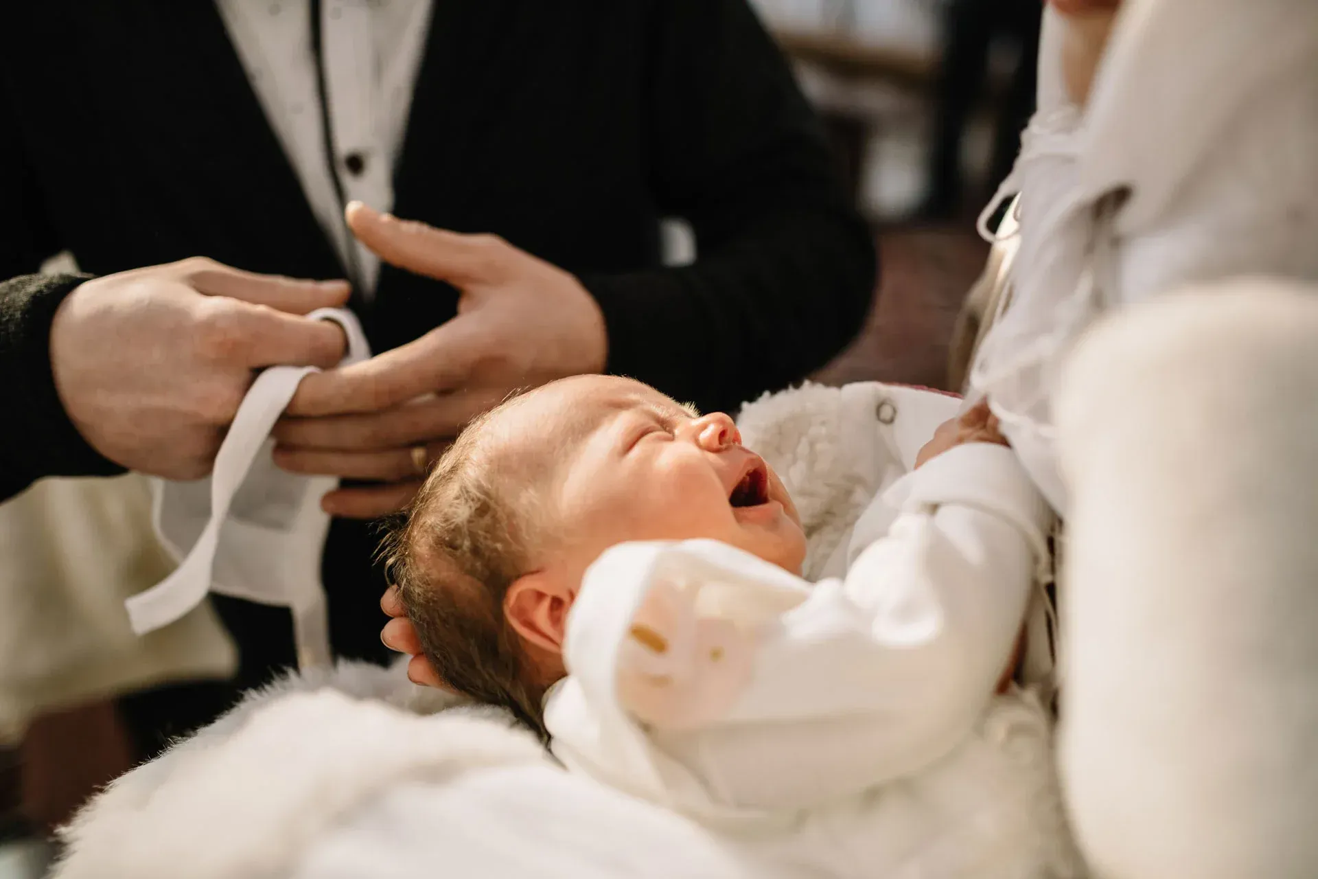 A baby cries during a baptism ceremony, held by a person wearing a white garment.