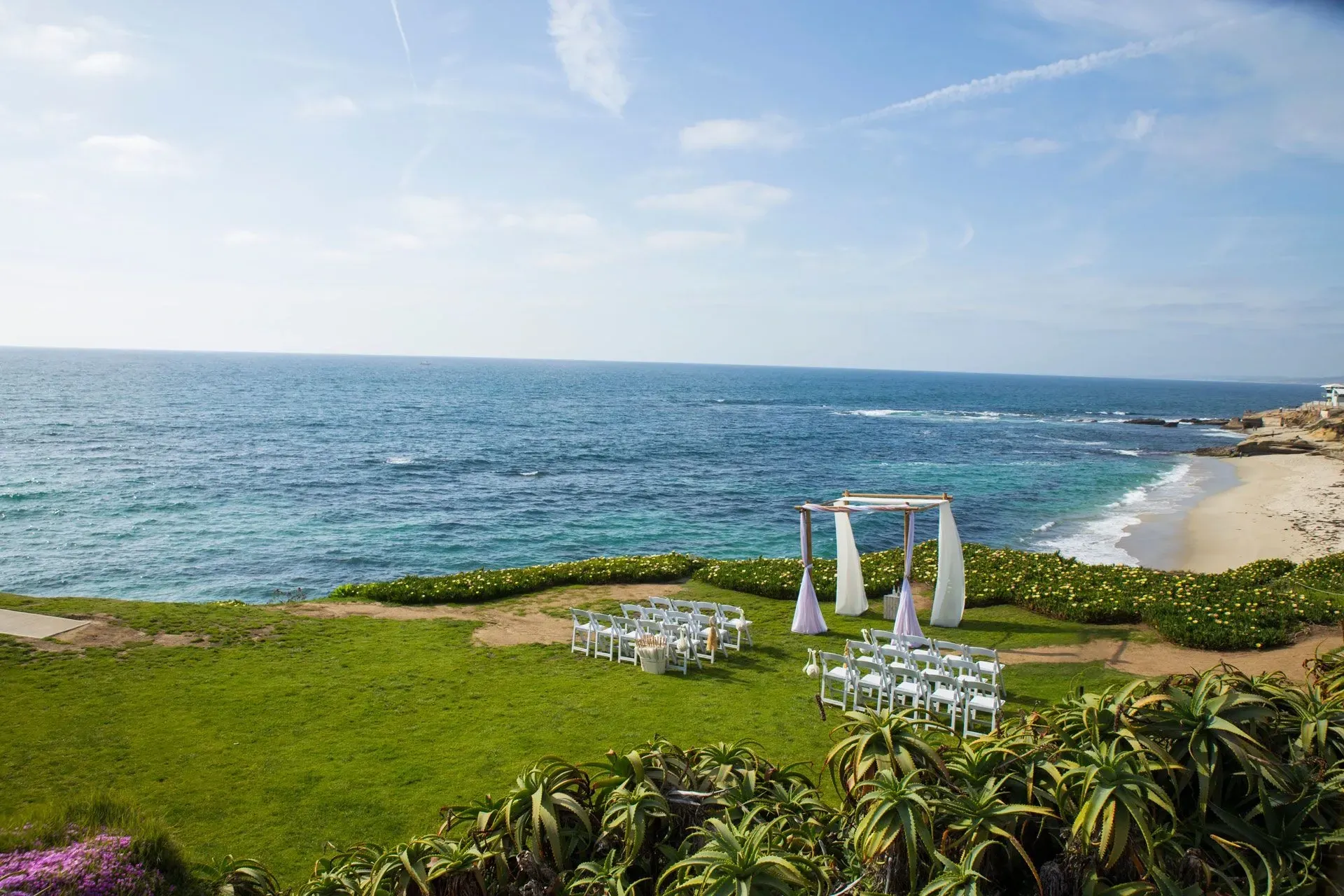 Wedding ceremony setup on a grassy cliff overlooking the ocean. White chairs, arch with fabric. Blue water and sky.