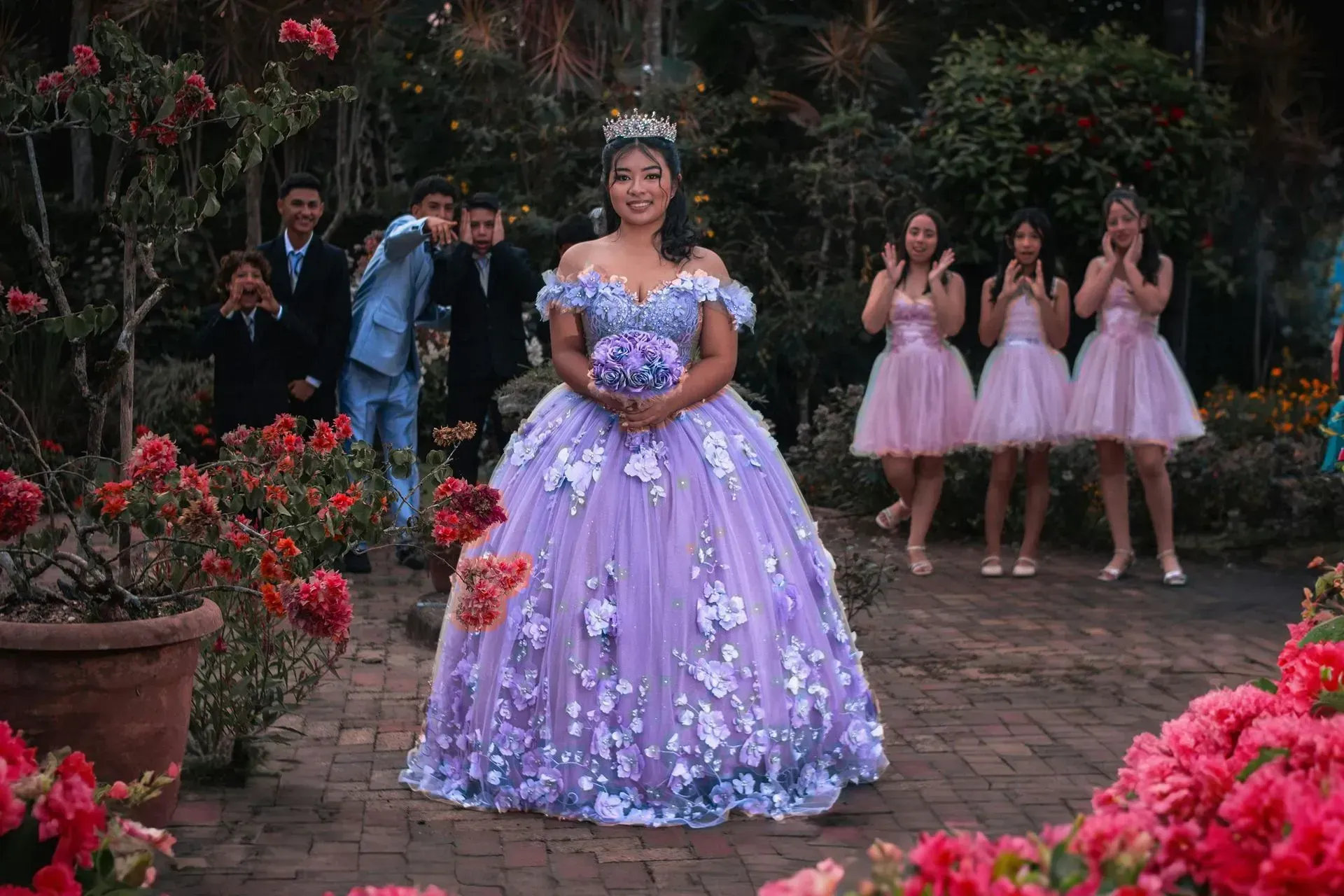 Girl in lavender ball gown with crown, holding bouquet, surrounded by bridesmaids and men in a garden.