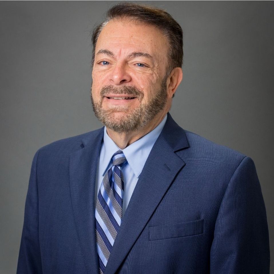 Man with a beard wearing a blue suit and tie smiles against a grey background.