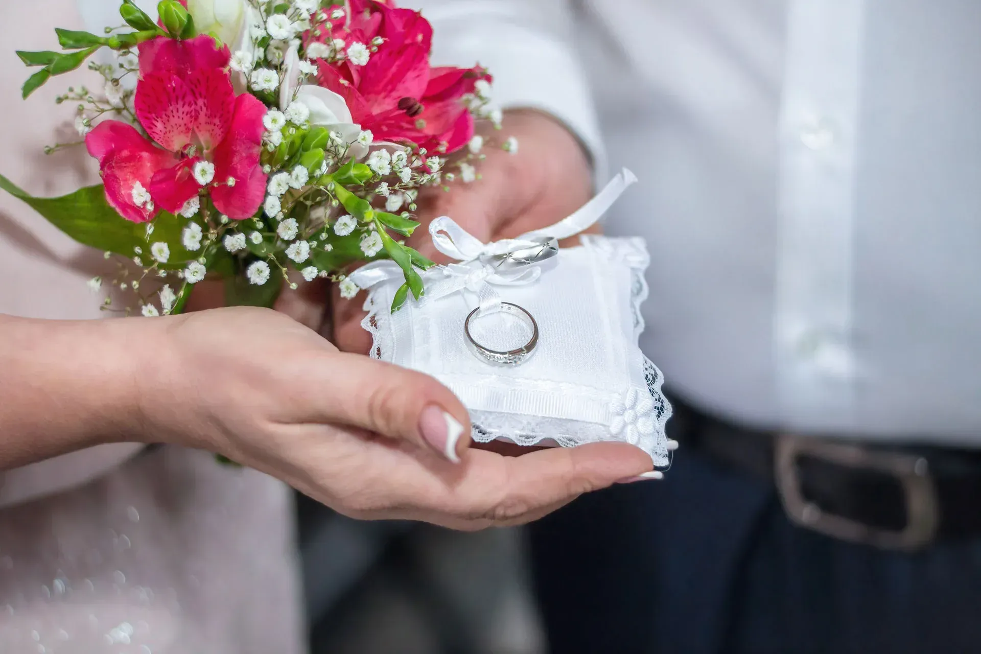 Hands holding a ring pillow with wedding rings, flowers in the background; formal attire.