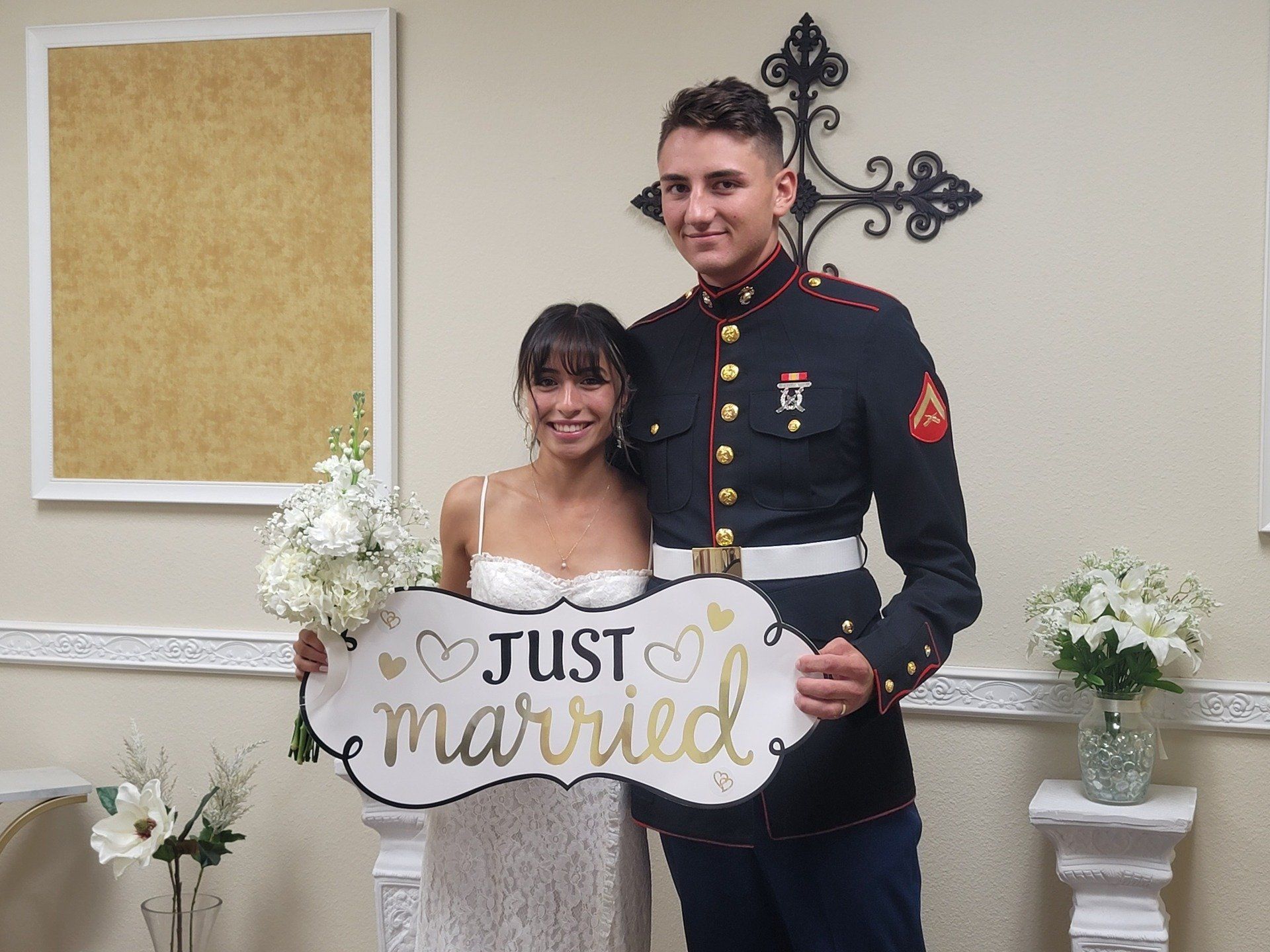 A Marine Groom and His Bride hold a Just Married sign in front of a Cross after their religious wedding