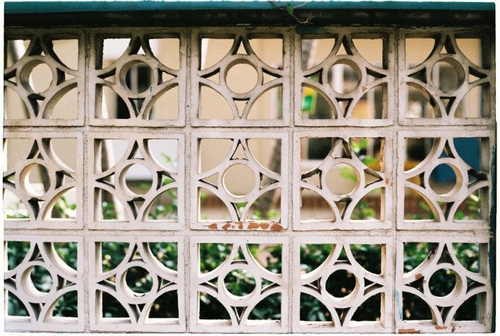 Decorative concrete wall with repetitive circular and geometric cutouts, revealing blurred greenery behind. 