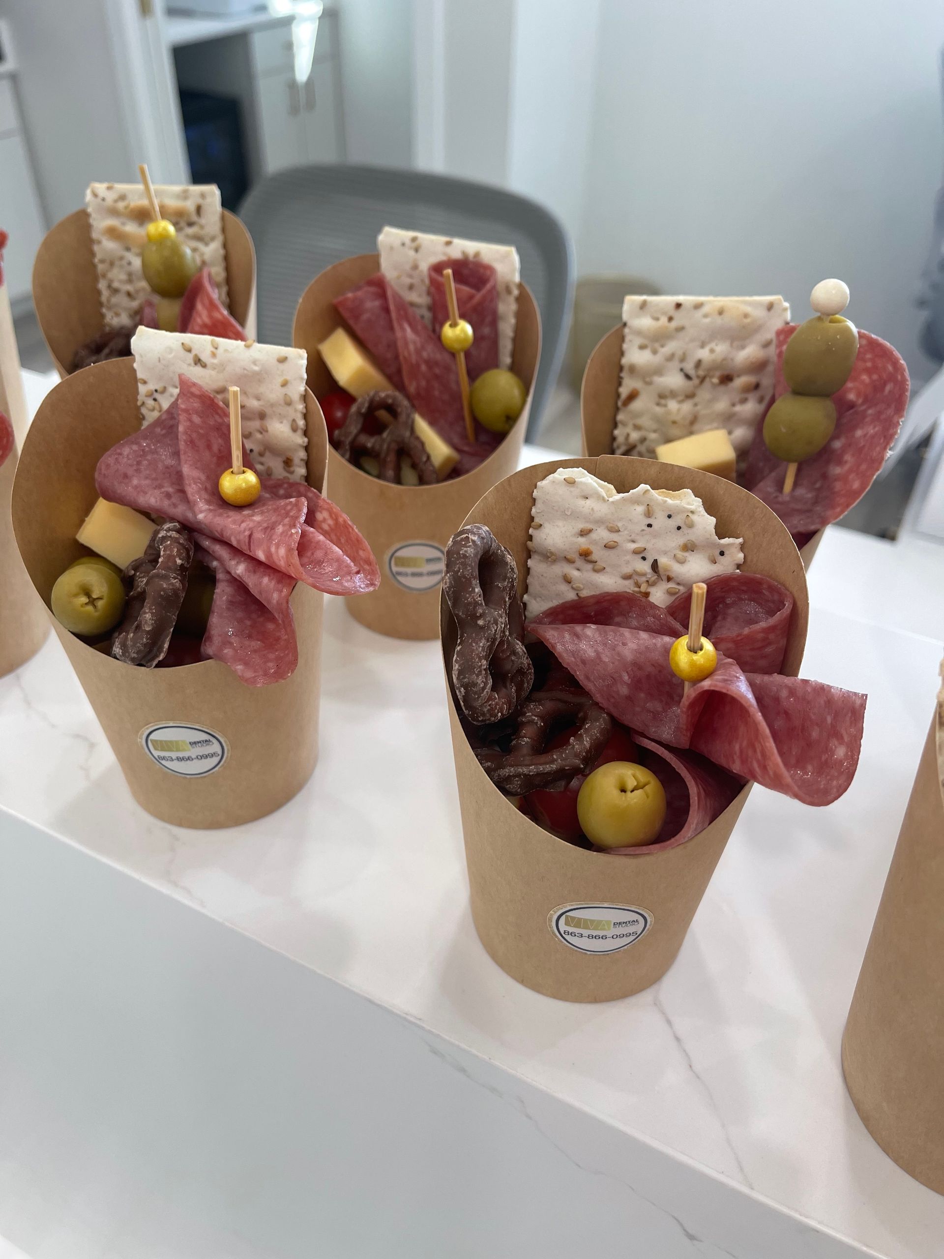 Charcuterie cups on a white surface: salami, cheese, olives, crackers, and pretzels in brown paper cones.