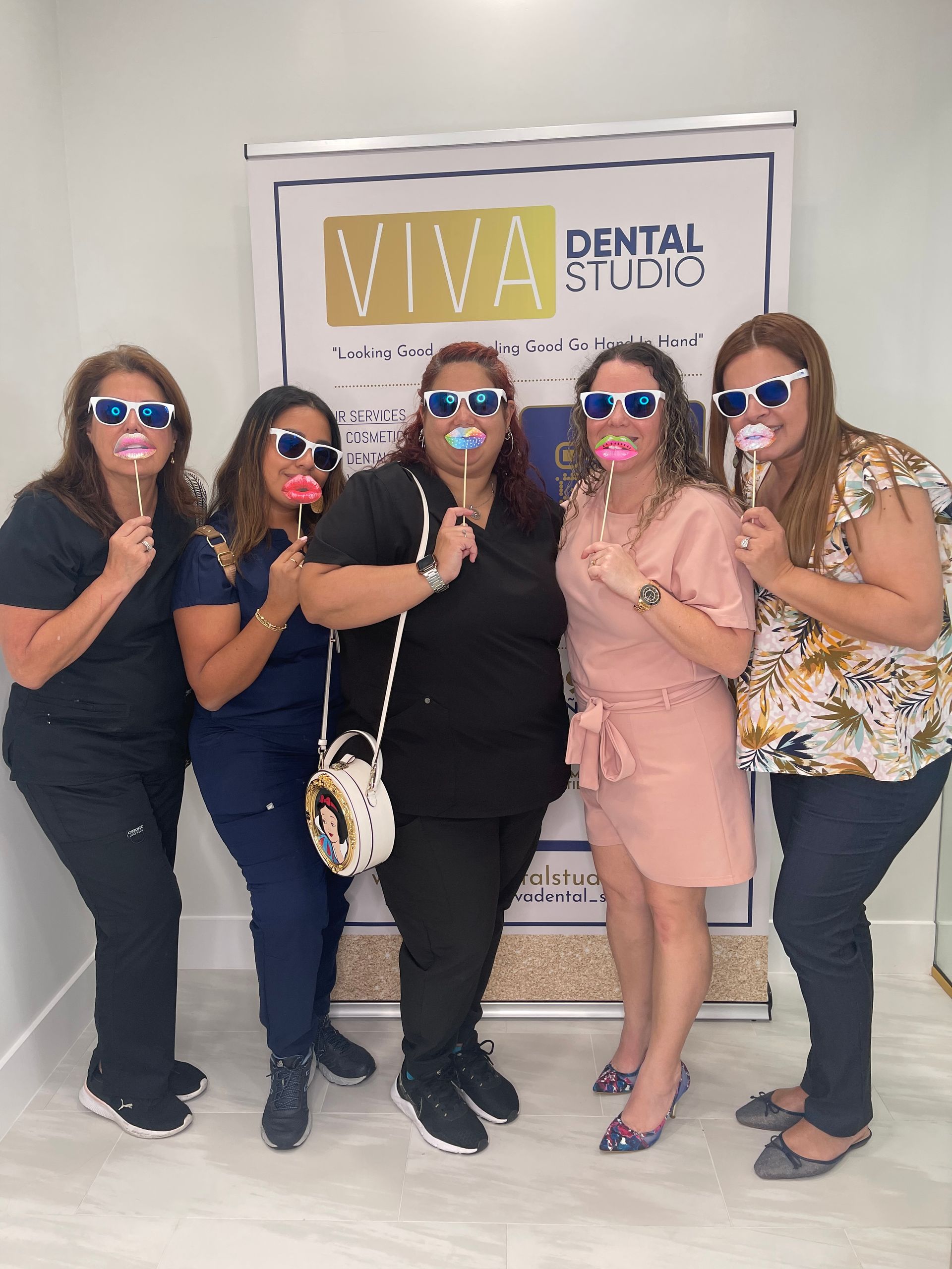 Five women holding prop lips and glasses posing in front of a dental studio banner.
