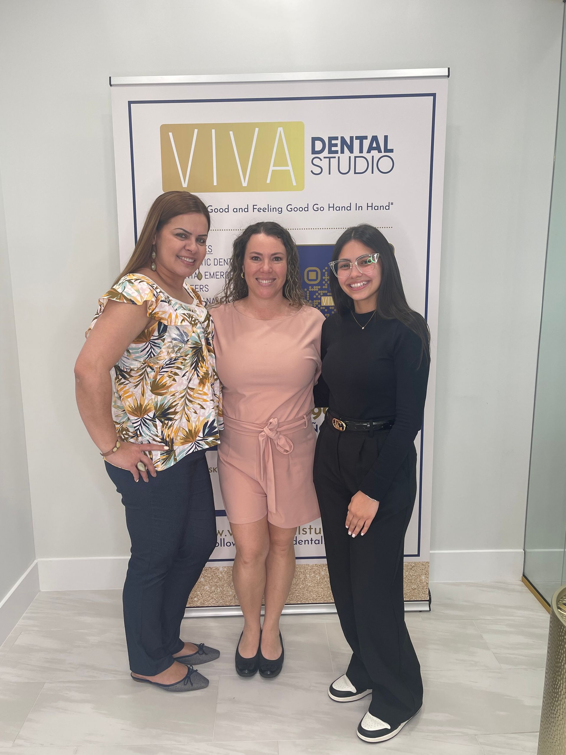 Three women stand in front of a dental studio banner. One wears a floral top, the other a pink dress, and the third a black outfit.