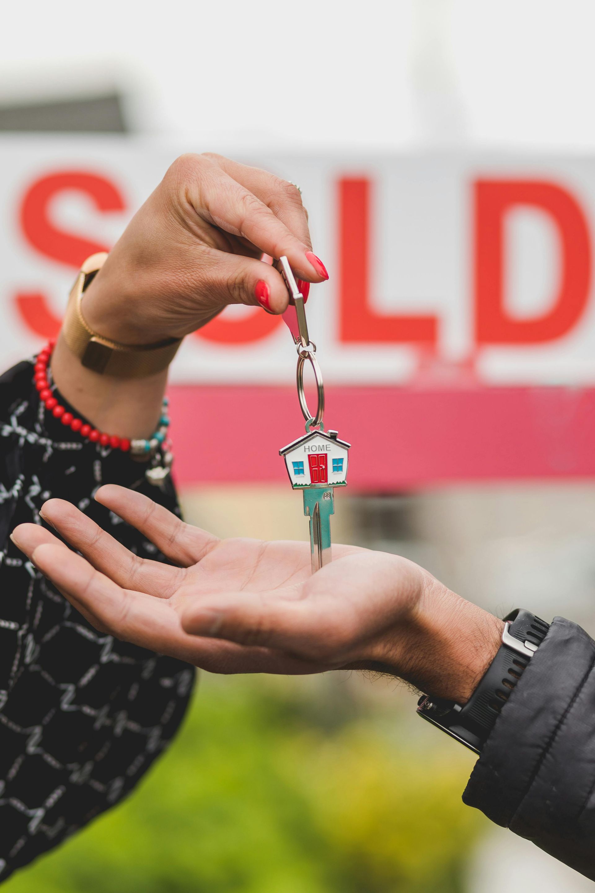 A person is handing a key to another person in front of a sold sign.